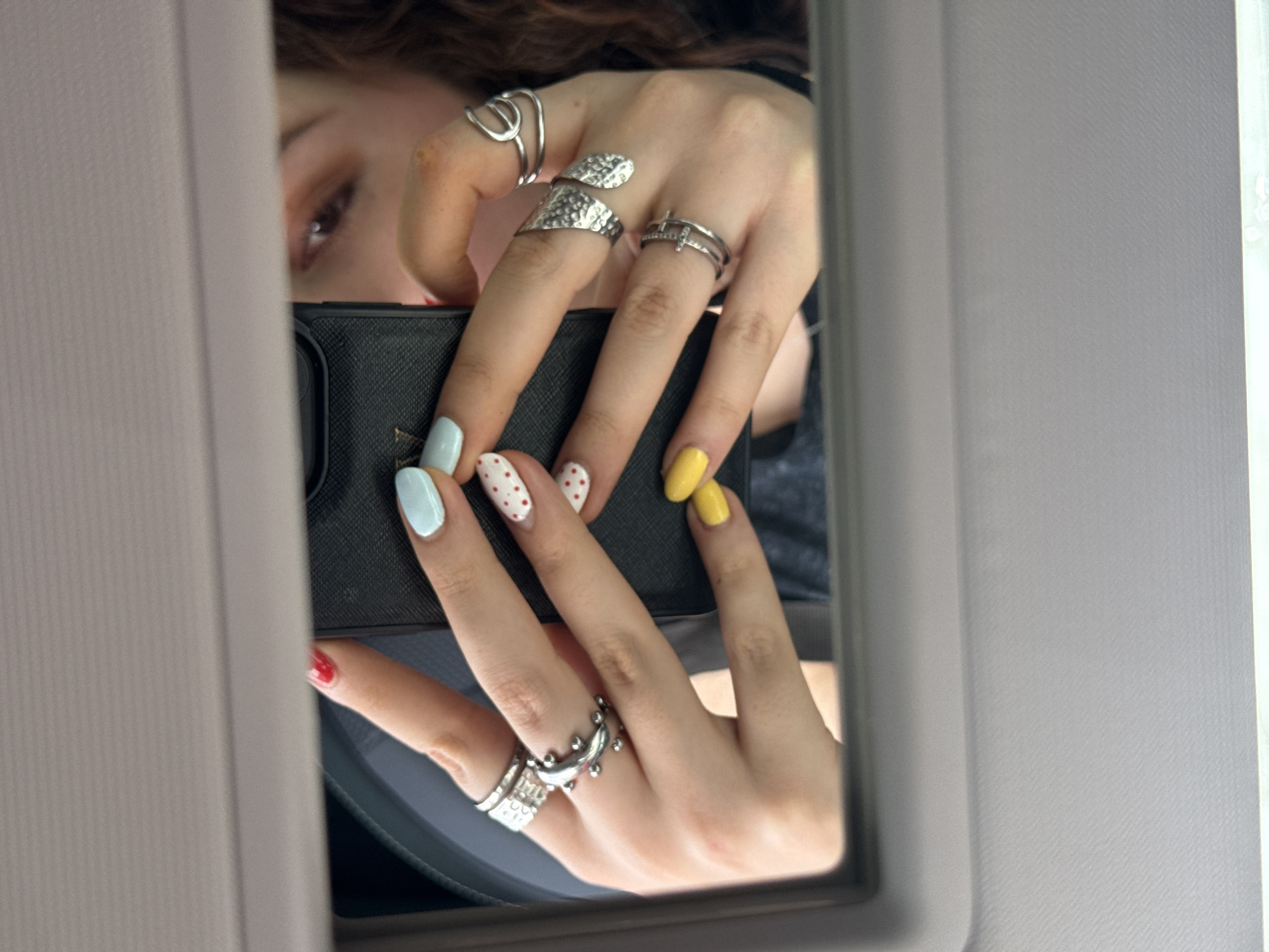 The image displays a close-up reflection of a person taking a selfie in a mirror, likely inside a vehicle. The upper part of the person's face is partially visible, showing a brown eye with makeup and brown hair peeking above a black smartphone.
The person's hands are the central focus, holding the smartphone in a selfie-taking pose. Both hands are adorned with multiple silver rings of various designs. On the visible fingers of the left hand, there is a large, textured silver ring on the middle finger, a thin double-banded ring on the ring finger, and an abstract, wavy ring on the index finger. The right hand features a ring with small, rounded protrusions on the ring finger and a textured band on the pinky finger.
The fingernails exhibit a playful and colorful manicure. On the left hand, the index and pinky fingers are painted light blue, while the middle and ring fingers feature white polish with red polka dots. On the right hand, the index and middle fingers are bright yellow, and the pinky finger is red.
The mirror is framed by a light gray, textured material, characteristic of a car's interior paneling or sun visor, suggesting the setting is within a car. The image captures a casual, personal moment. No discernible text is visible on the phone or elsewhere in the reflection. While the location context is Mexico City, Mexico, the visual information in the image itself does not provide specific cues to confirm this location beyond the general car interior setting.