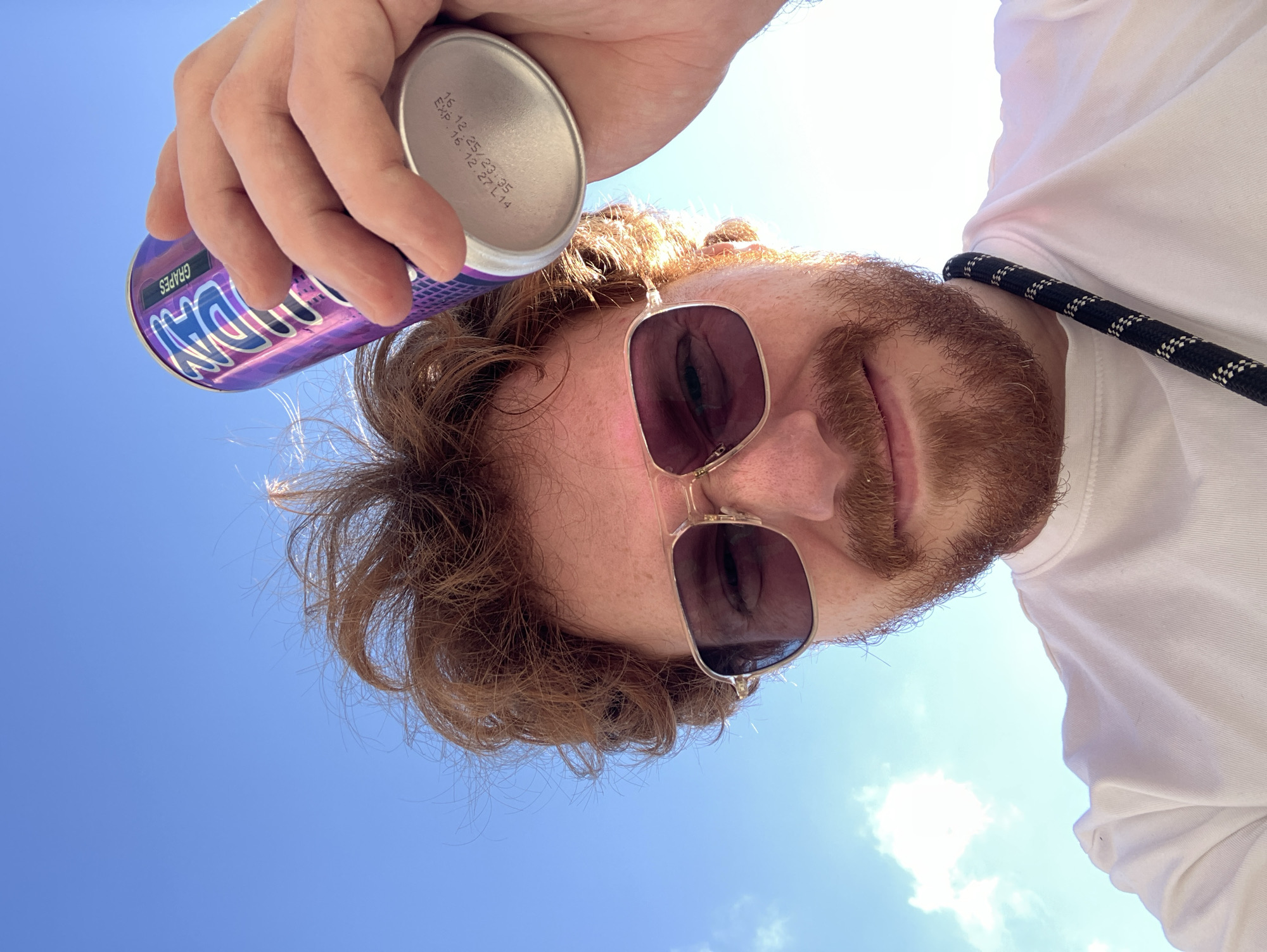 A low-angle shot captures a man against a bright blue sky with scattered white clouds, suggesting a sunny outdoor setting in Tel-Aviv, Israel. The man has reddish-brown hair and a matching beard. He wears dark, gold-framed sunglasses and a white shirt, with a black-and-white patterned lanyard or strap visible near his shoulder. He looks towards the camera with a slight, friendly smile.
He holds a purple soda can in his left hand, positioned above his head. The can's label clearly shows the brand "DAN" and the flavor "GRAPES", along with a small number "654395". The bottom of the can displays manufacturing or expiration details, including "16.12.25/23:35" and "EXP:16.12.27L14". The bright daylight illuminates his hair and face, creating a relaxed, casual atmosphere on a clear day.