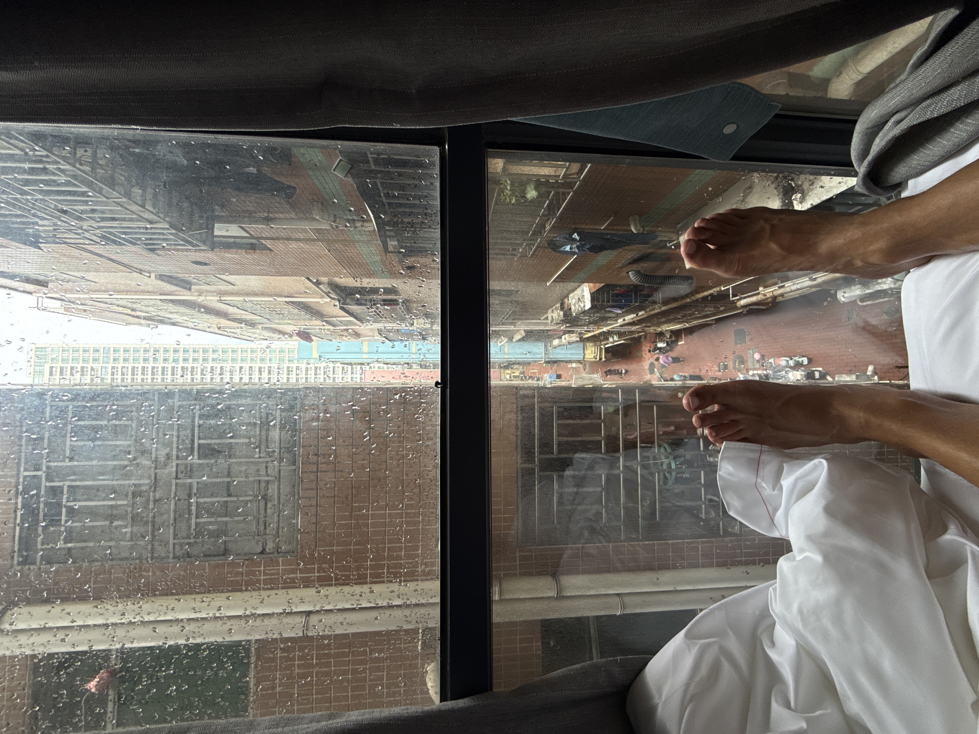 The image captures a view from inside a building in Futian District, China, looking out through a rain-streaked window. In the foreground, a pair of human feet are visible, resting on white sheets in what appears to be a bed. The window offers a glimpse of the exterior environment, showcasing adjacent buildings and a street scene below. The weather is rainy, indicated by the water droplets on the windowpane. The image is oriented at an angle, giving an upward perspective of the buildings across the street. The scene is framed by the room's interior, with portions of the wall and possibly a headboard or bed frame visible.