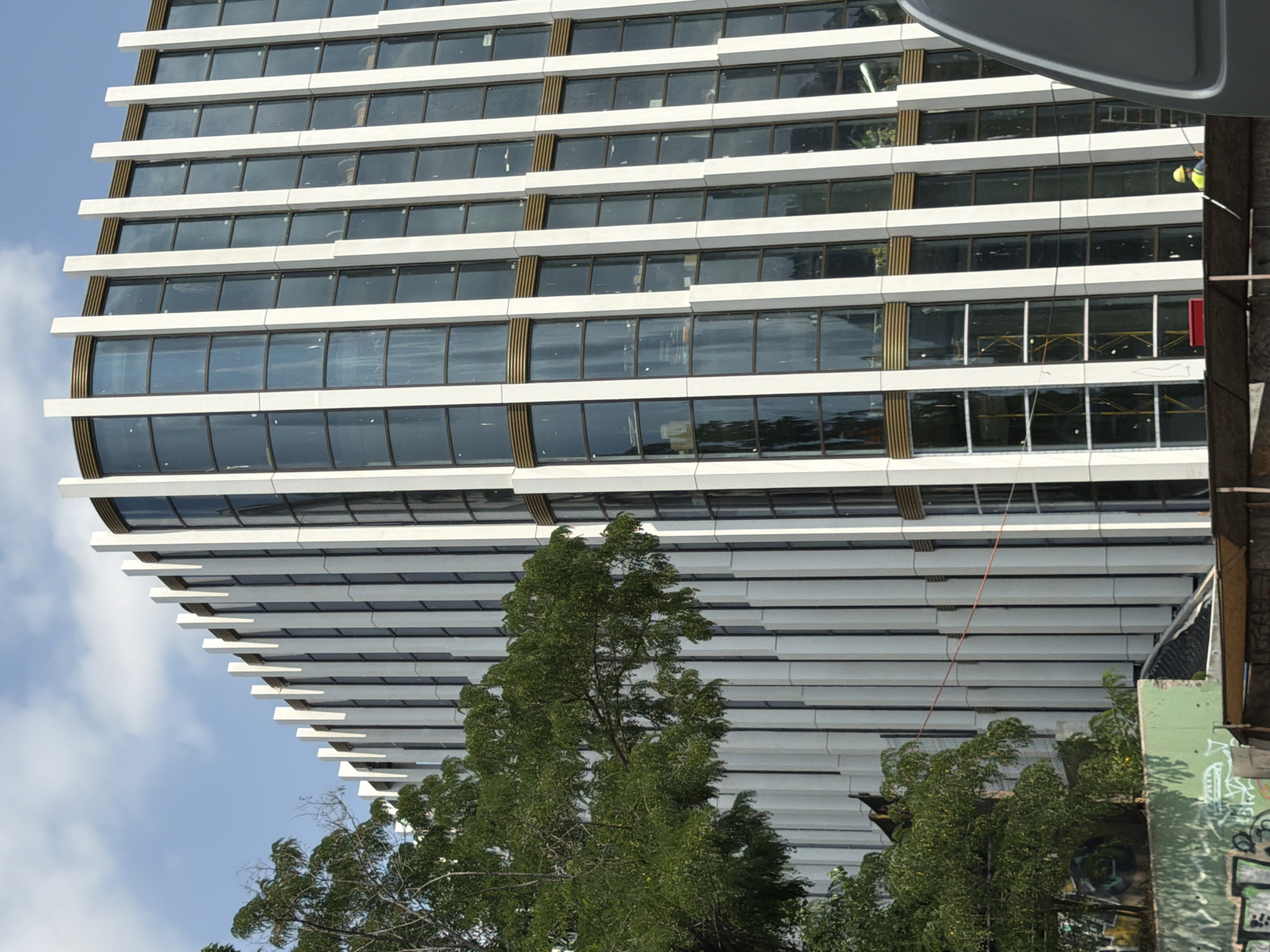 A medium shot captures the exterior of a modern, white-paneled building under construction in Tbilisi, Georgia. The building features vertical white fins that divide large, dark-tinted windows, some of which are curved. A thin gold band accents the building horizontally between some window sections. To the left, a large, leafy green tree partially obscures the building's facade. In the foreground, a section of a green wall covered in graffiti is visible, along with scaffolding and construction materials. A construction worker wearing a yellow hard hat and a blue safety vest is visible near the bottom right, holding a pole. The sky above is blue with scattered white clouds.