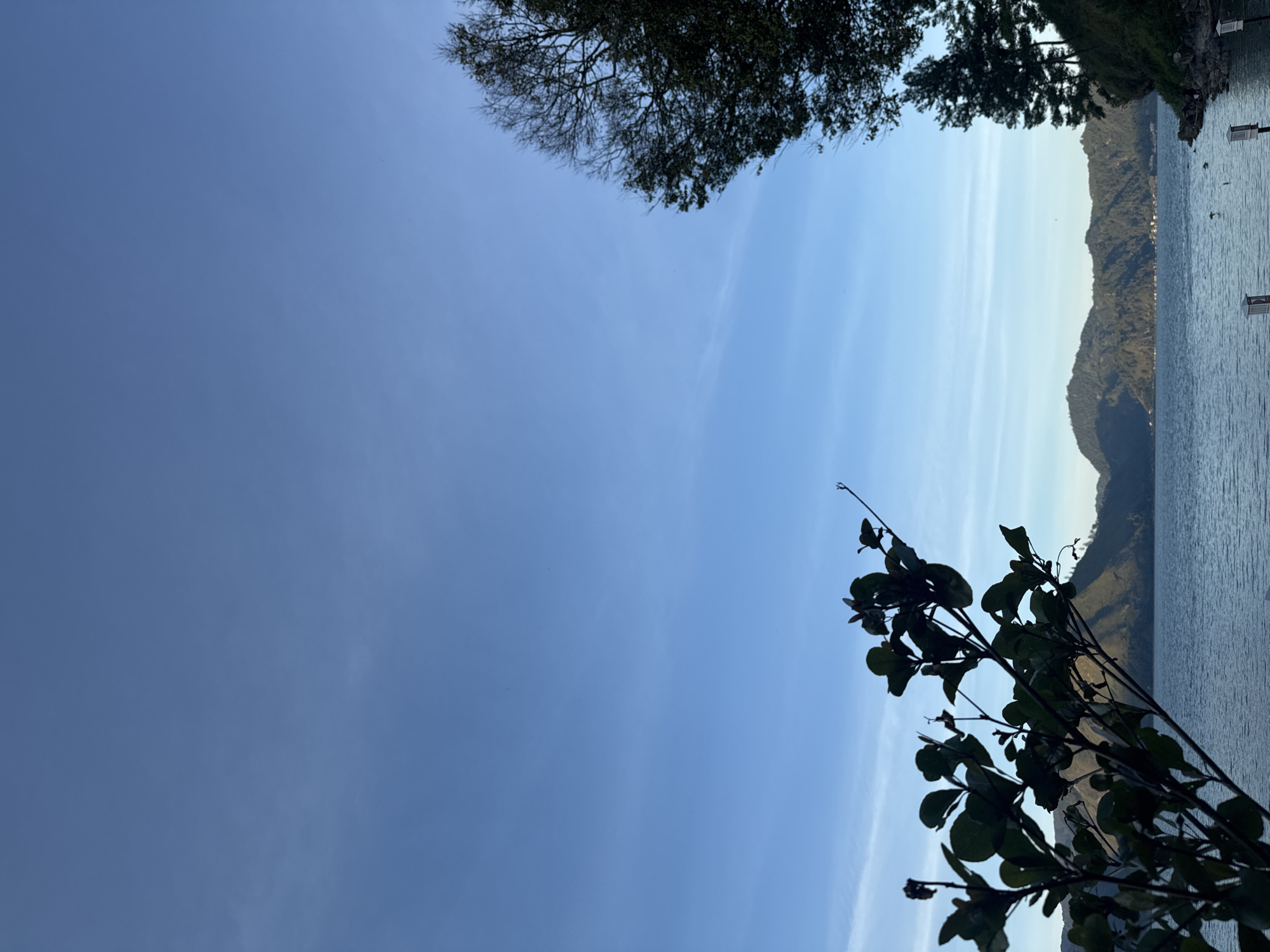 This is a landscape photograph of Lake Toba in Kalalengkio, Indonesia. The image is dominated by a clear blue sky with wispy white clouds. In the foreground, a dark silhouette of a leafy branch extends from the left side of the frame. To the right, a portion of a bare-branched tree and several evergreen trees are visible. In the middle ground, the calm blue water of Lake Toba stretches across the frame, bordered by a range of lush green mountains. A few small buildings are clustered along the base of the mountains. The overall scene suggests a serene and natural environment.