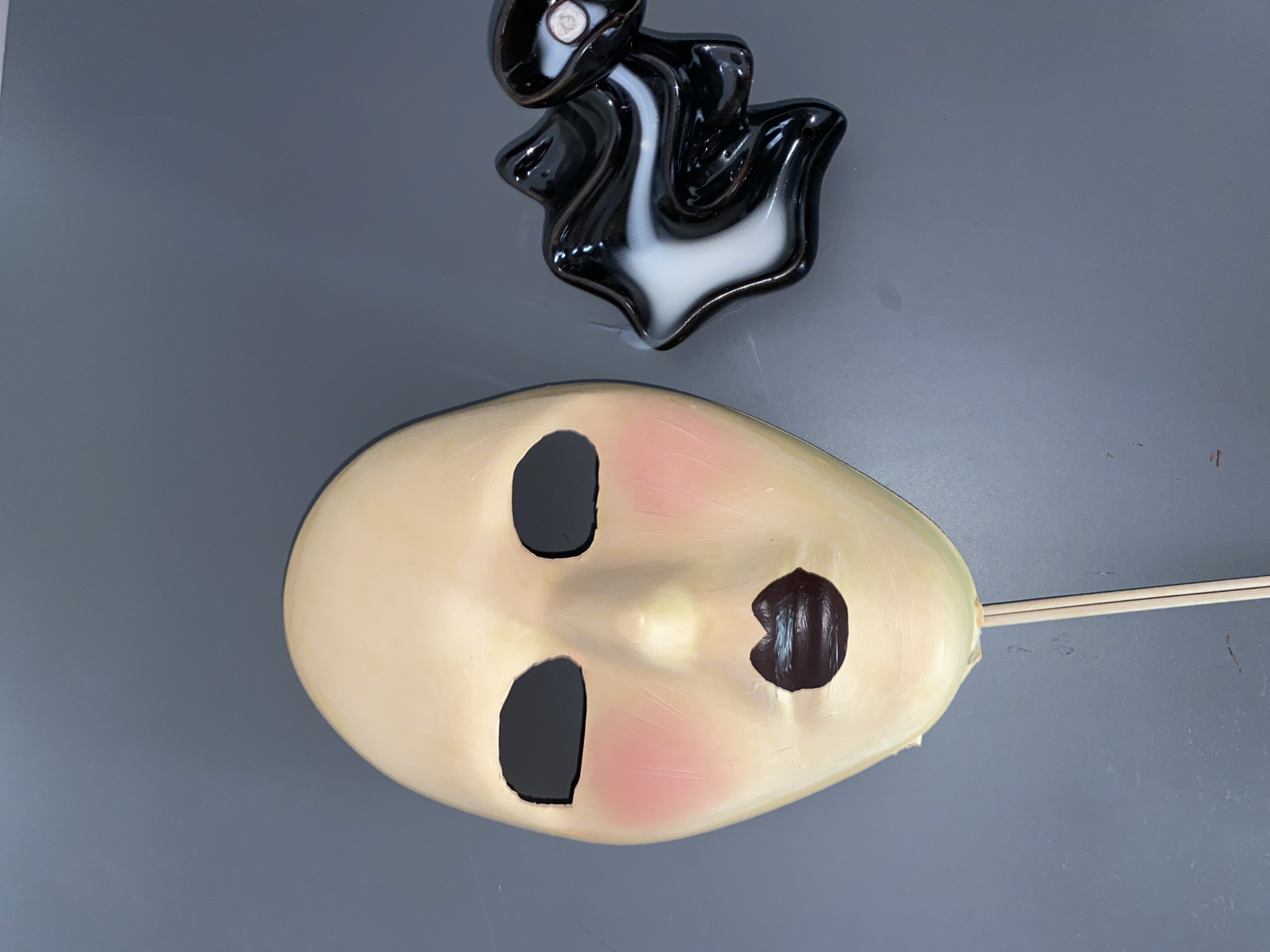 An oval mask-like object with vacant eye sockets and a button-like feature on the right side lies on a gray surface. The mask is flesh-toned with reddish patches on the cheeks. A black and white decorative piece is above the mask. A thin, light-colored stick extends from the right side of the mask. The location is not discernible.