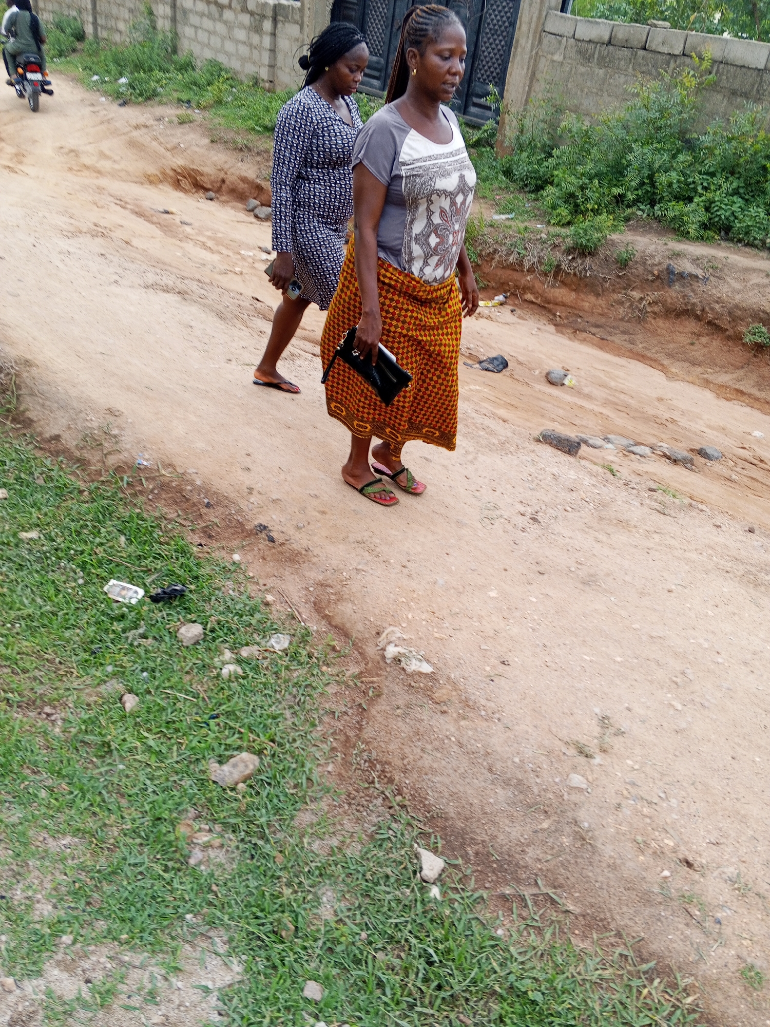 Two women walk along a dirt road in Zango-ndaji, Nigeria. One woman wears a patterned blue dress, while the other wears a gray t-shirt and an orange and yellow patterned wrap. The second woman carries a black clutch. Litter is visible on the roadside. In the background, a person rides a motorcycle. A concrete block wall and green vegetation are also visible.
