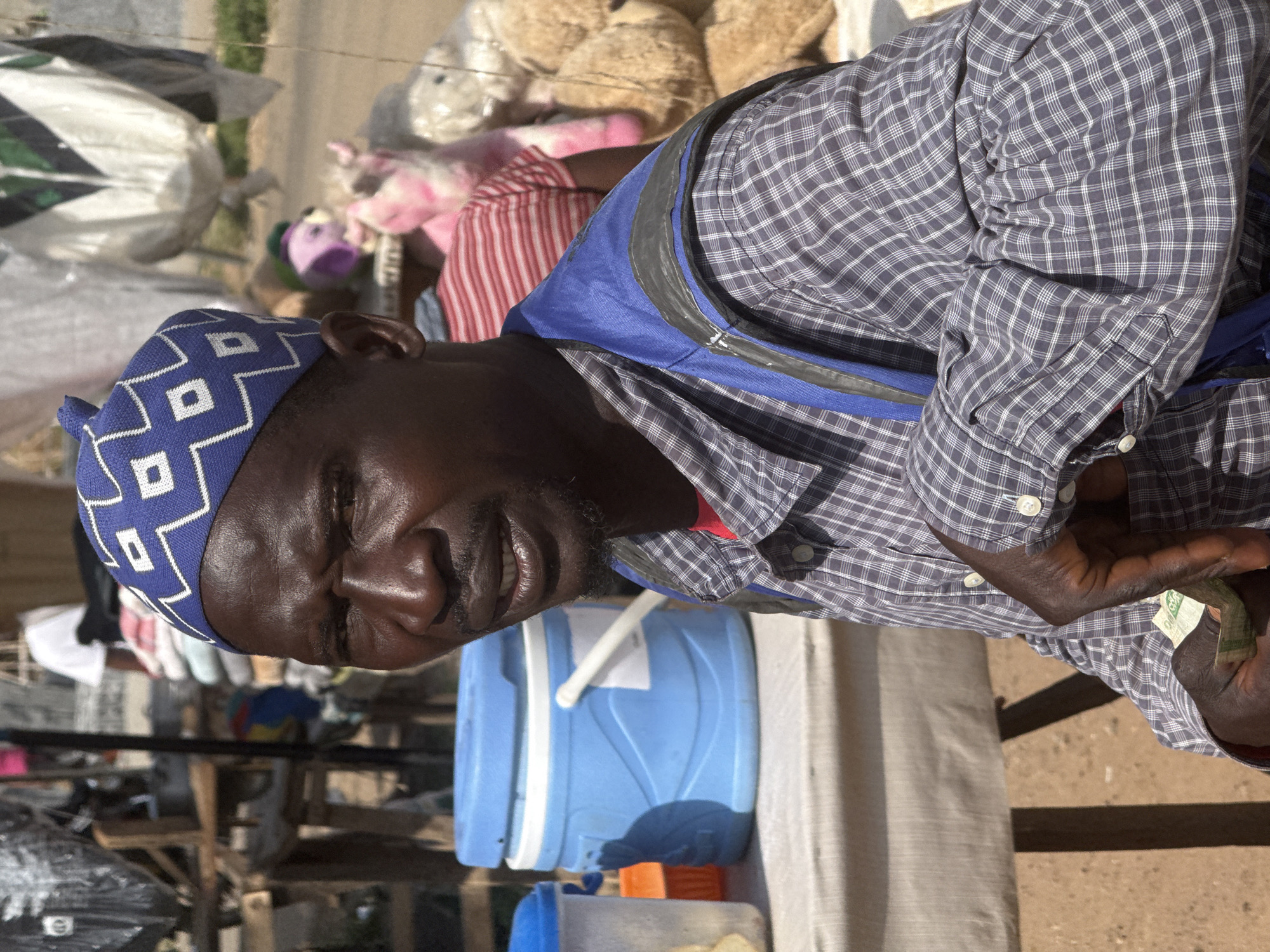 The image portrays a dark-skinned man standing in an outdoor, market-like setting in Abuja, Nigeria, under bright daytime light. The man, possibly a vendor, is angled towards the viewer with partially closed or squinting eyes and a subtle, relaxed smile, suggesting either enjoyment or a reaction to strong sunlight.
He wears a blue head covering adorned with a white geometric pattern, a blue and grey plaid long-sleeved shirt, and a blue utility vest or backpack with black accents over his shirt. In his left hand, he holds several folded green and white currency notes, one of which clearly displays "100", identifying them as Nigerian Naira.
To the man's right, a light blue plastic water cooler or dispenser with a white lid and handle sits on a light-colored surface, possibly a stall or table. A smaller blue container is visible beneath it. The background is bustling with market activity, featuring piles of items including plush toys (pink and brown) and large white and patterned bags. The ground appears to be dirt, reinforcing the outdoor, informal commercial environment. The sharp highlights and shadows indicate a sunny day.