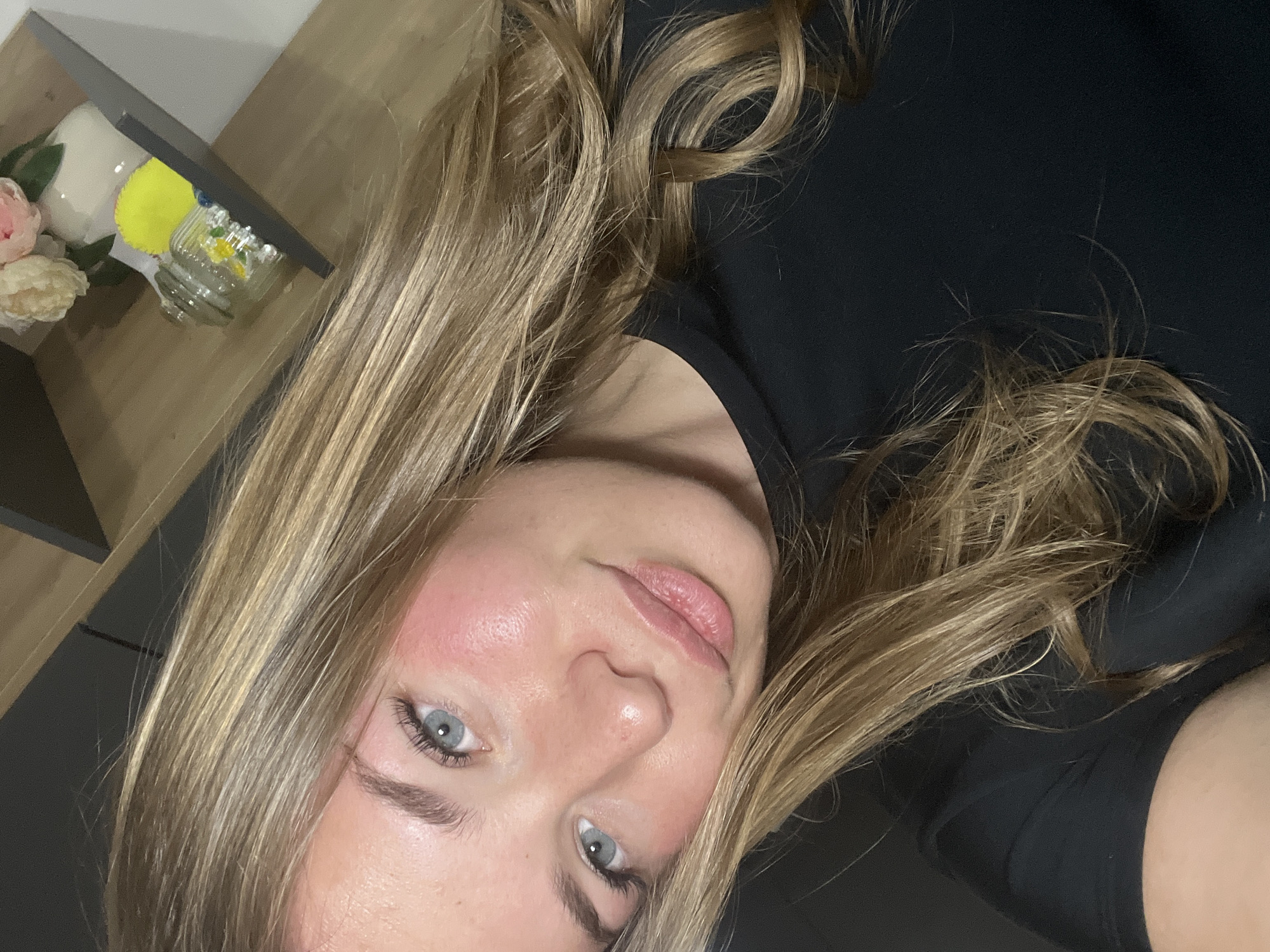 A close-up, slightly angled selfie shows a light-skinned woman with long, blonde hair and light blue eyes. She is wearing a black top. In the upper left corner of the frame, a shelf is visible with decorative items, including flowers and a small jar. The city of Budva, Montenegro is not discernible from the visual information provided in the image.