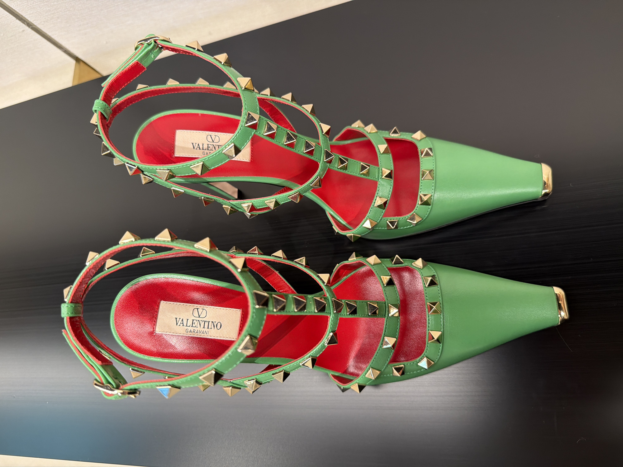 A pair of green Valentino Garavani studded heeled sandals are displayed on a black surface. The sandals are bright green with red insoles and feature multiple straps adorned with gold pyramid studs. A gold buckle is visible on the ankle strap of the left sandal. The background is dark with a subtle reflection. This image was taken in New York City, United States of America.