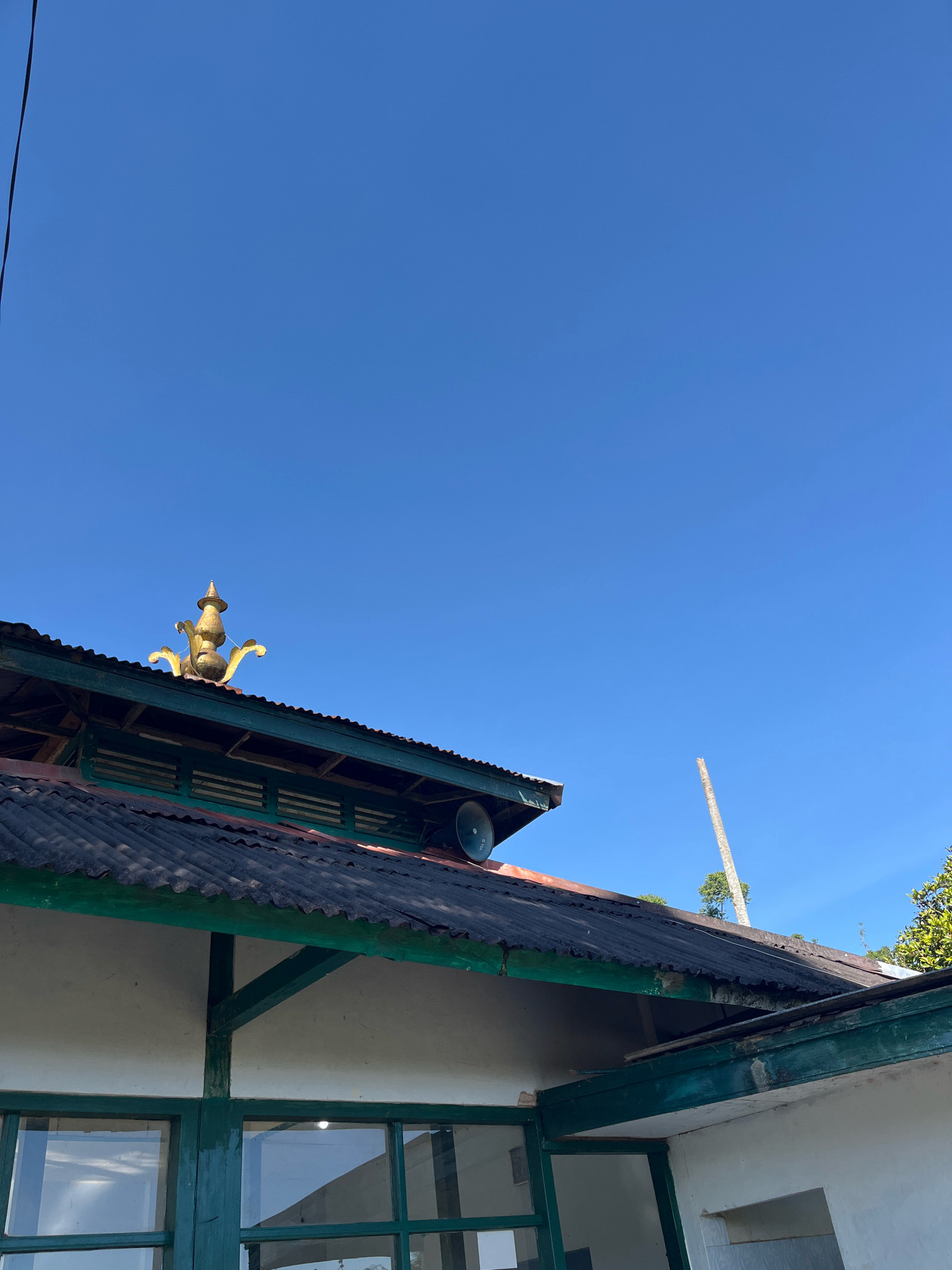 The image shows the roof and upper exterior of a building under a clear blue sky. The roof is made of corrugated metal, some of which appears weathered and aged. A decorative golden finial, resembling a lamp or spire, is visible on the peak of one section of the roof. There is also a speaker mounted on the side of the roof. The building's exterior walls are white, with green trim around the windows and roofline. Some wooden beams are visible supporting the roof structure. In the background, a tall, slender tree or pole is visible against the sky, along with some green foliage on the right side of the image. The scene appears to be outdoors, possibly in a rural or semi-rural area. The location is identified as Tomblo, Indonesia.