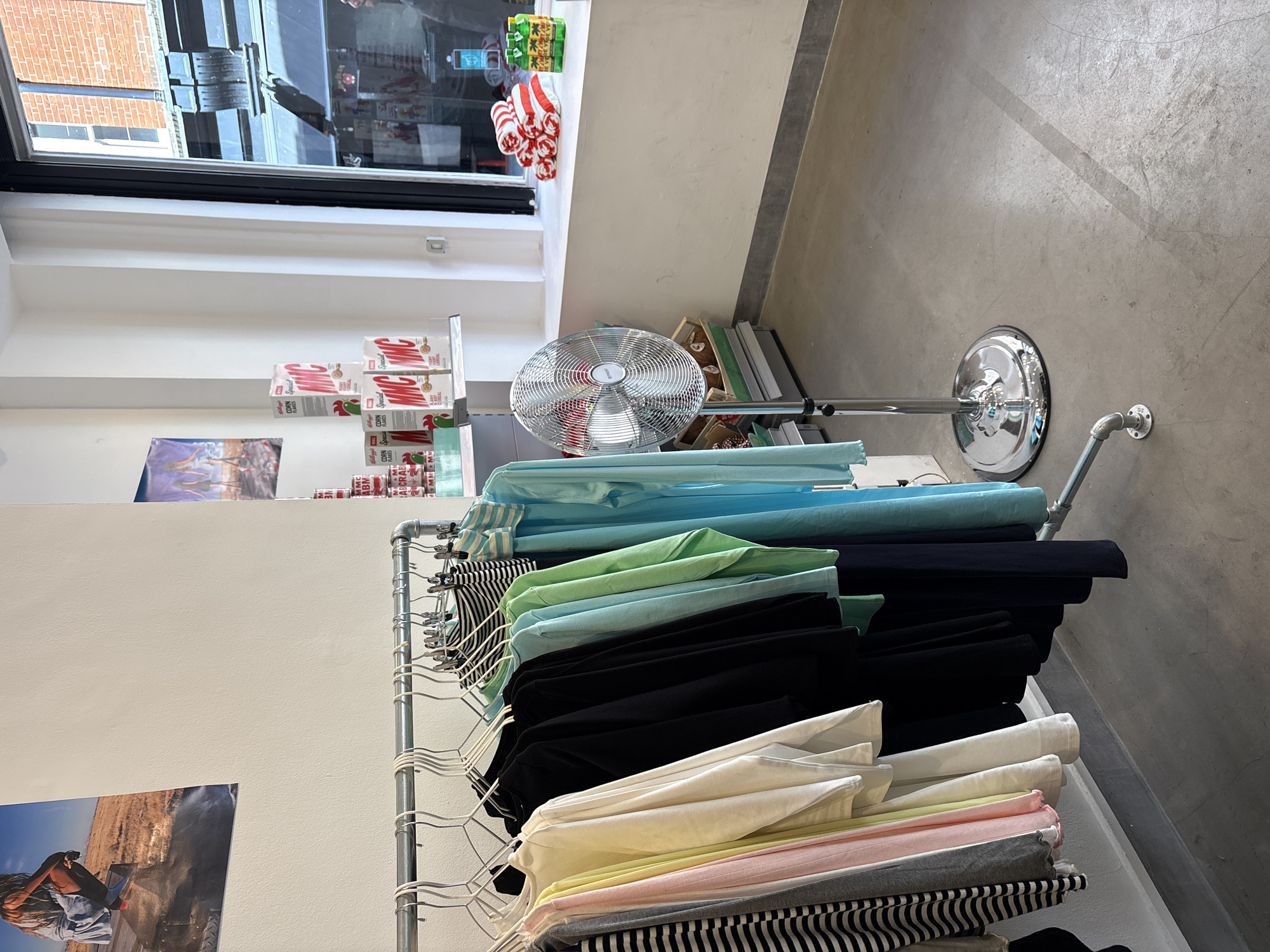 The scene is an interior space, possibly a clothing store or a boutique, in what appears to be London, United Kingdom. The image is taken at a high angle, looking down at a clothing rack displaying a range of garments. The clothing consists of multiple layers, with colors transitioning from light blue at the top to green, black, white, pink, grey, and striped patterns at the bottom. A metal fan on a chrome stand is visible in the background. The store's window, showing a brick building across the street, indicates that the image was taken during daylight hours, with natural light coming in through the window. The foreground shows a poster featuring an image of a person sitting by a canyon or a cliff. The background contains boxes on a shelf. The overall atmosphere is clean and well-lit, giving off the impression of a modern retail environment. The flooring is polished concrete.