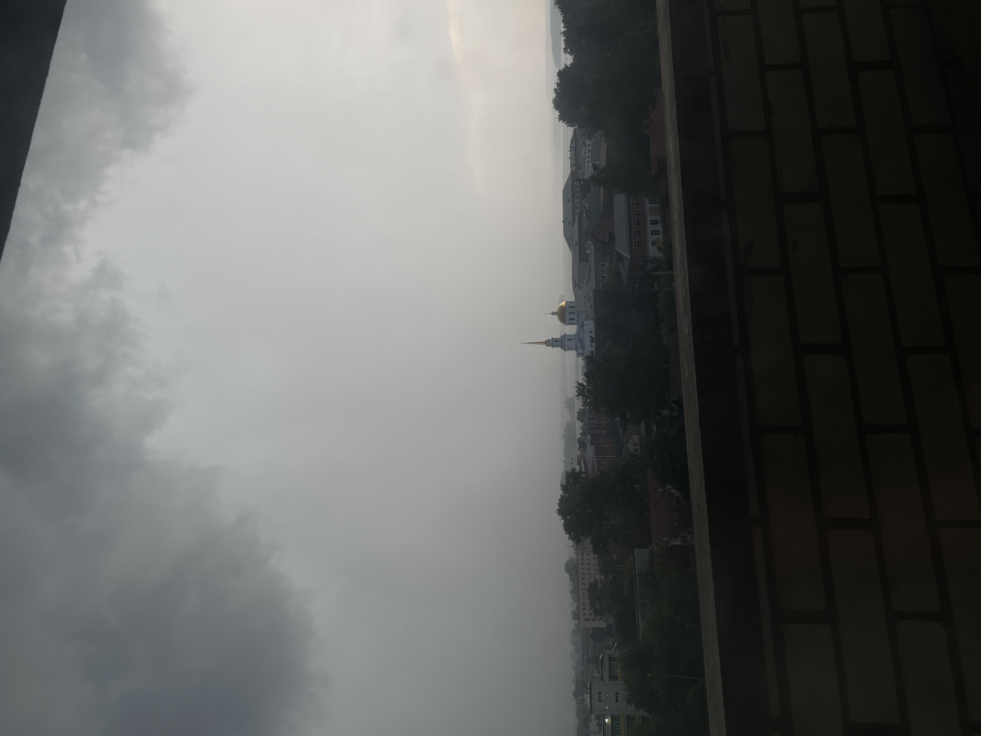The image depicts a view of Votkinsk, Russia, from a high vantage point. The scene is dominated by a thick layer of overcast clouds, suggesting a potentially stormy or hazy day. In the distance, the outline of buildings and trees can be seen through the mist, with a prominent golden-domed structure, likely a church, standing out against the muted sky. The city appears to be situated along a body of water, possibly a river or lake, hinted at by the distant horizon.
The foreground is partially obscured by a dark, textured surface, appearing to be a brick wall or a railing, which frames the view of the city. The composition is rotated, with the sky and city appearing on the left side and the textured surface on the right. The lighting is subdued, consistent with a cloudy day, and there is no indication of any specific activity or event taking place, other than the general presence of a cityscape. The overall mood is somewhat somber and atmospheric due to the heavy cloud cover. No text is visible in the image.