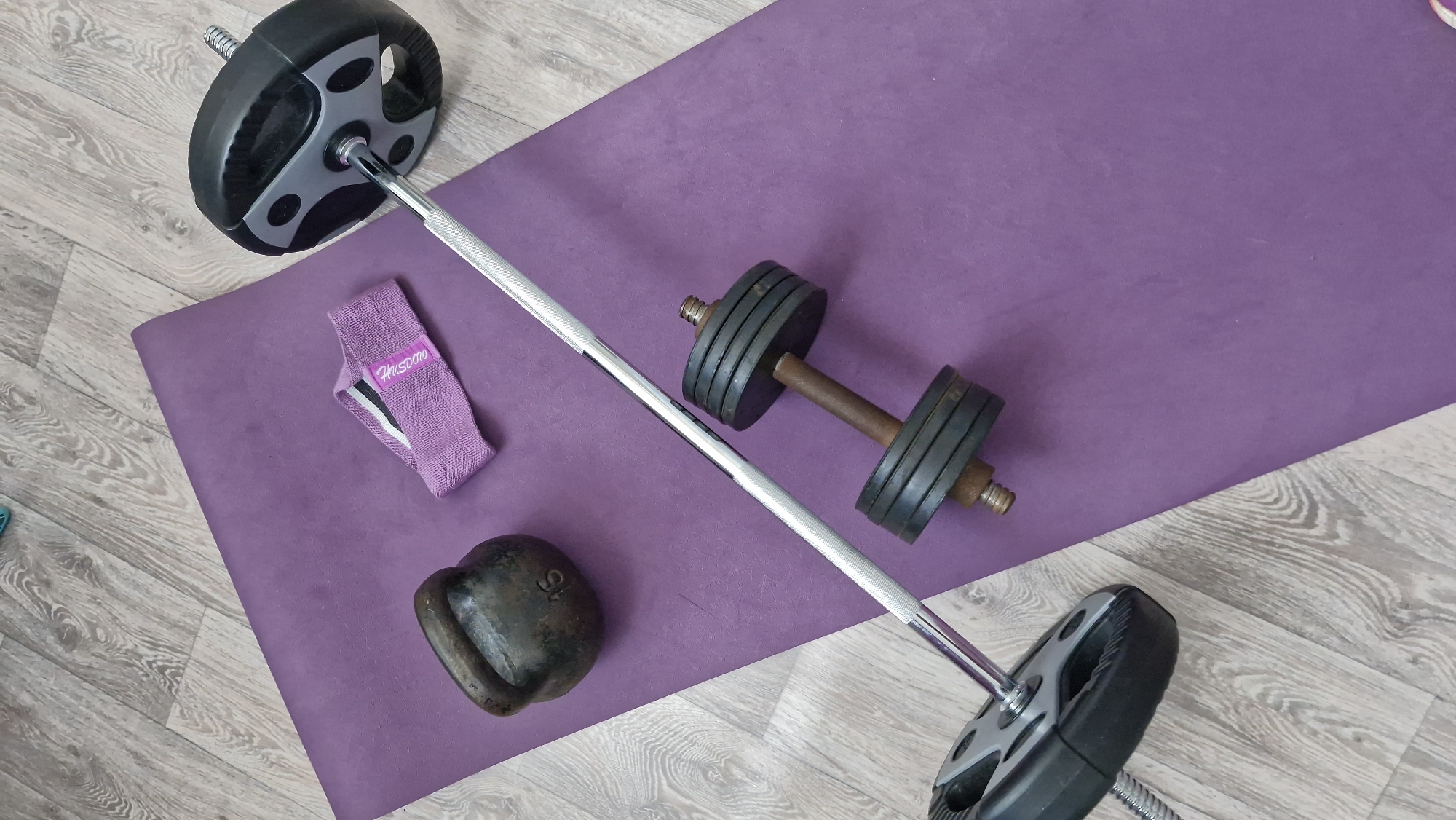 A high-angle shot shows a purple yoga mat on a light wooden floor, upon which a barbell with black and gray weights on either end lays diagonally. To the left of the barbell are a purple resistance band with the word "HUSKDOV" printed on it and a black kettlebell with the number "16" visible on its side. To the right of the barbell is a dumbbell with a rusty, brown handle and four black weights on either end. The scene is indoors, possibly in a home or fitness studio.