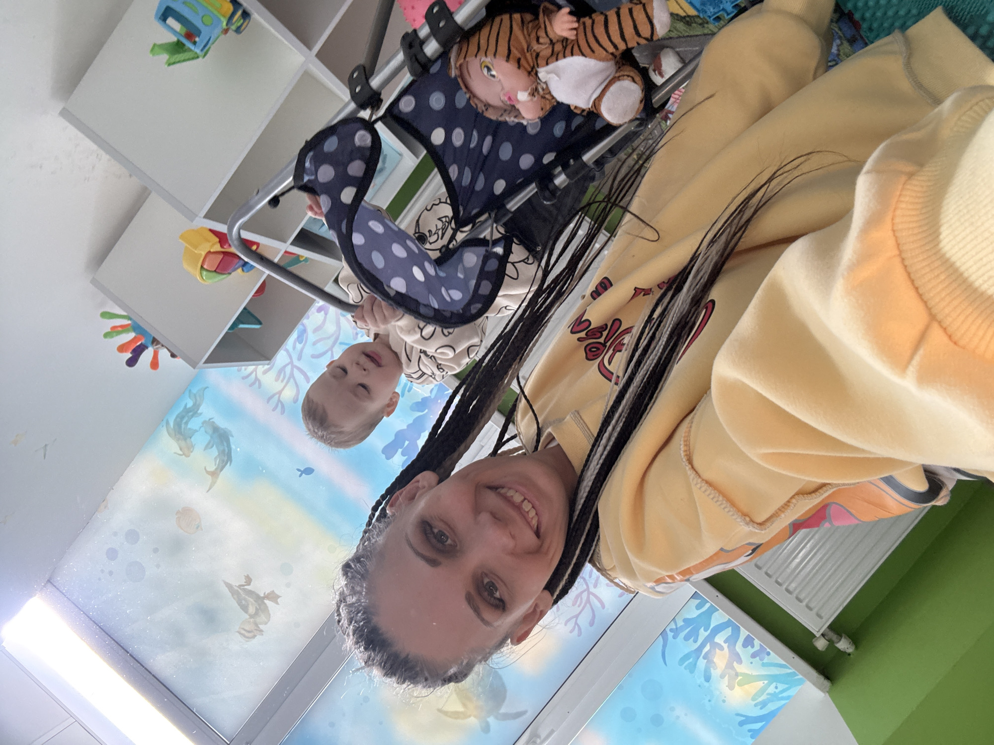 The image captures a woman and a child in a room, likely in Rusanovo, Russia, though precise location confirmation would necessitate additional details. The woman is smiling directly at the camera, with braided hair. The child is seated in a stroller alongside a tiger-striped doll. The room features a shelf with toys, a wall painted green, and a ceiling with a mural of an underwater scene.