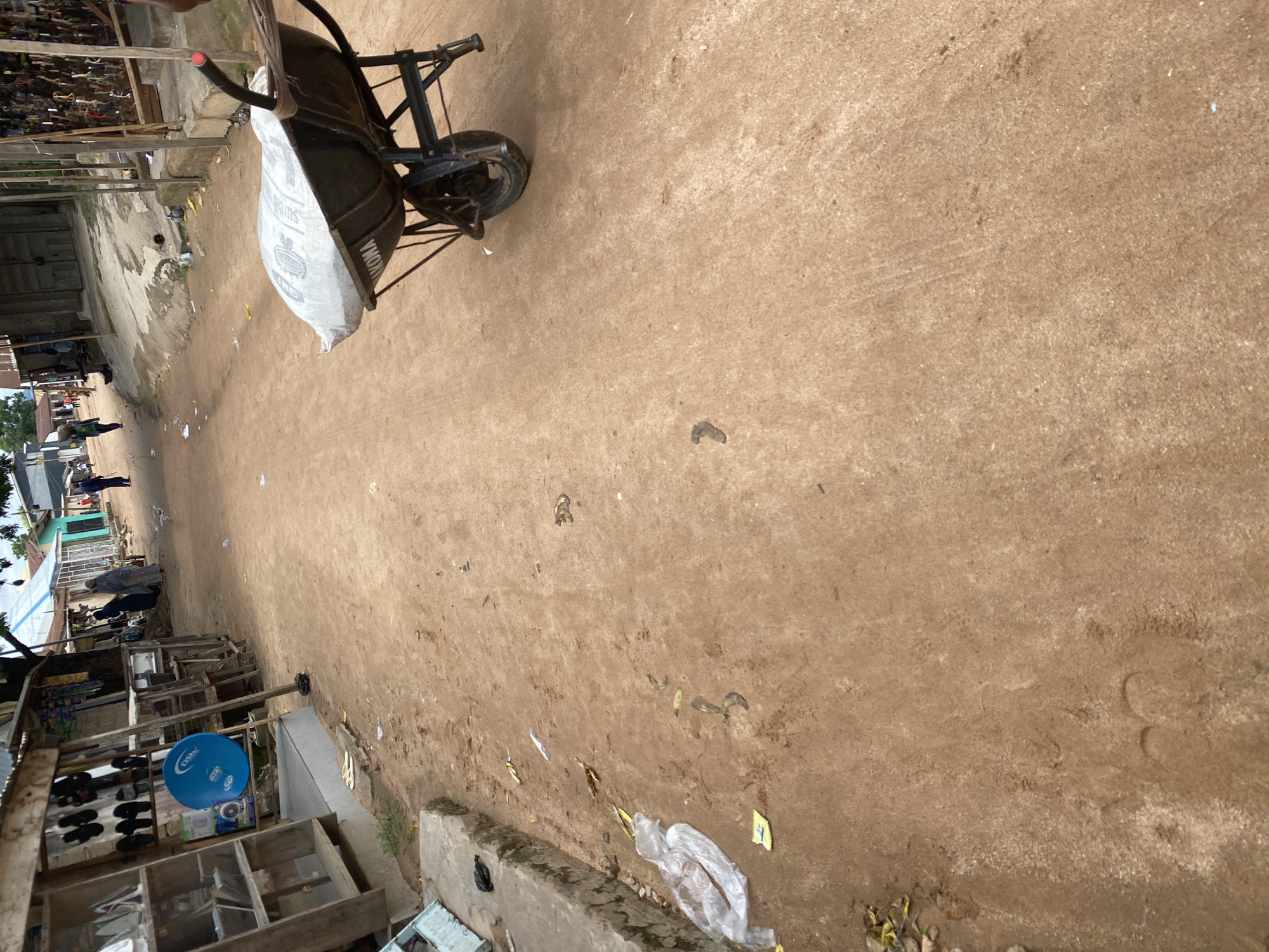 The image depicts a street scene in Jalingo, Nigeria, with a dirt road that appears to be a thoroughfare. On the left side of the road, a stall or shop is visible, featuring shelves stocked with various items, including footwear. A blue satellite dish is also mounted on a pole. The road itself is unpaved and shows signs of wear and tear, with scattered debris, including a white plastic bag and what appear to be banana peels.
In the middle ground of the image, a wheelbarrow is positioned, loaded with a large white sack. The wheelbarrow's handles are visible, and one handle has a red marker or tape on it. Beyond the wheelbarrow, several individuals are walking along the road. One person in the distance is carrying a load on their head. Further down the road, there are more buildings and structures, some with corrugated iron roofs. The overall environment suggests a local market or a residential area with commercial activity. The time of day appears to be daytime, with natural light illuminating the scene.