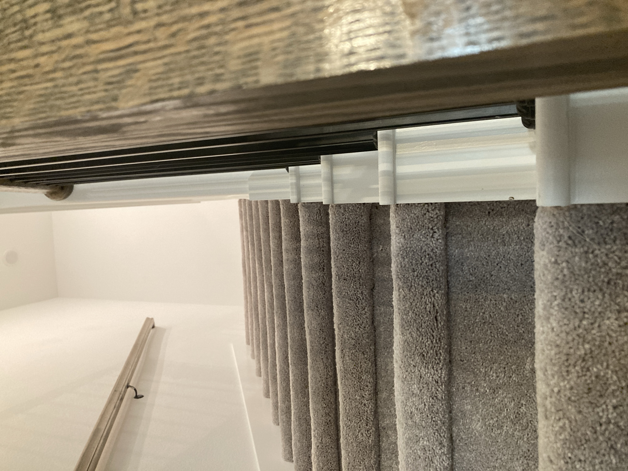 The image shows a close-up, upward-angle view of a carpeted staircase in what appears to be a residential interior. The staircase is covered in a light gray, textured carpet. To the right of the stairs, a white balustrade with decorative detailing is visible, alongside black metal spindles and a wooden handrail. On the left side of the frame, a wooden handrail is mounted on a white wall. The environment is well-lit, suggesting an indoor setting, likely a home or apartment building. There are no people or animals visible in the image. The location is not specified, but the style of the staircase and balustrade suggests a modern residential setting. This scene is consistent with residential interiors found in Round Rock, Texas, United States of America.