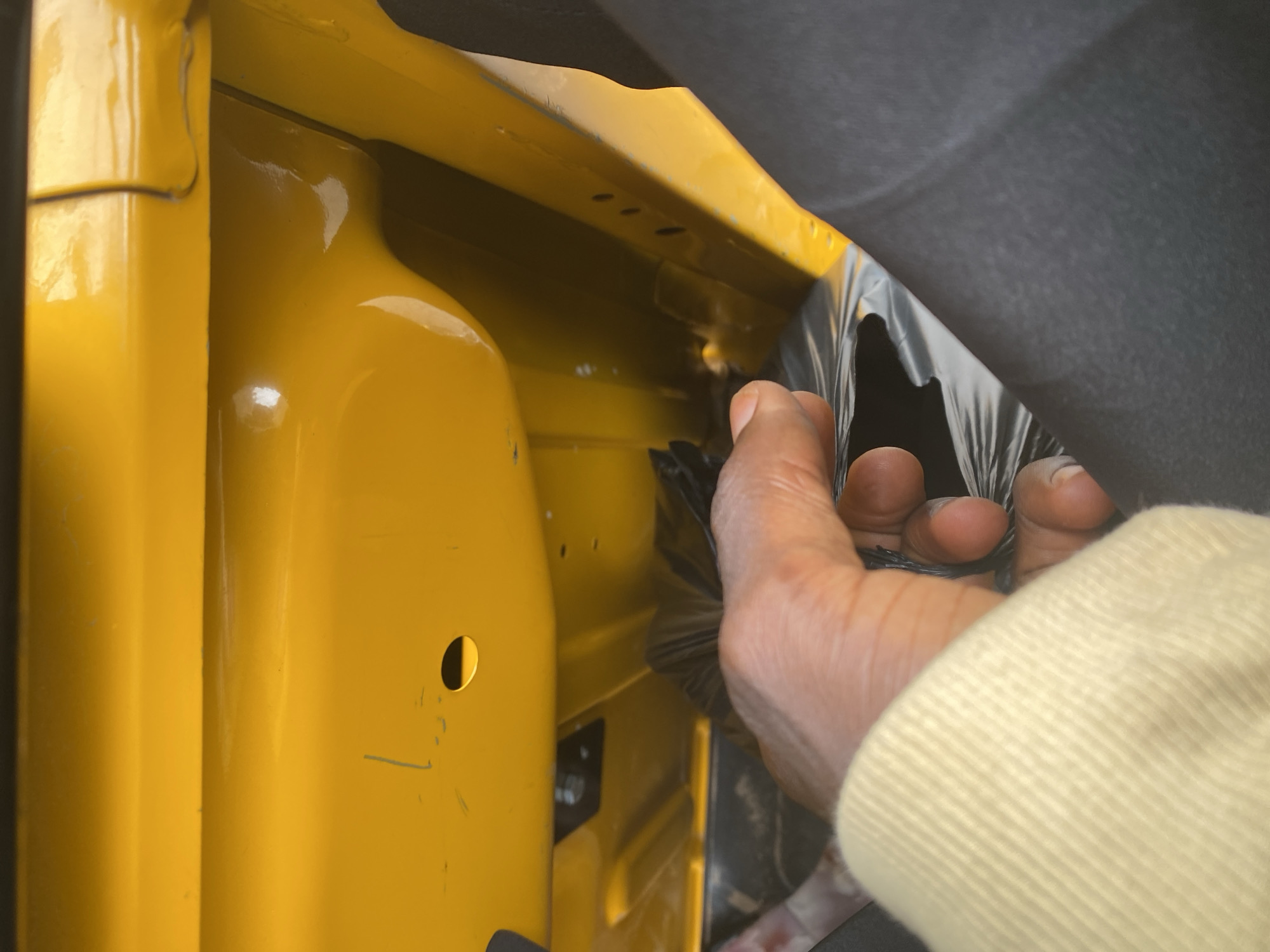 A person's hand is visible, reaching into a yellow object, possibly the interior of a vehicle. The hand is holding a black, crinkled material, which appears to be a plastic bag or sheeting. The yellow object has a smooth, glossy surface and features some indentations and a small circular hole. Part of a dark gray or black material is also visible, possibly upholstery or another interior component. The lighting suggests it might be daytime, with bright reflections on the yellow surface indicating a well-lit environment. No text is visible in the image. The location context of Jalingo, Nigeria, provides a geographical setting but does not offer specific visual cues within the image itself. The overall impression is a close-up view of someone interacting with an object, potentially for storage or organization.