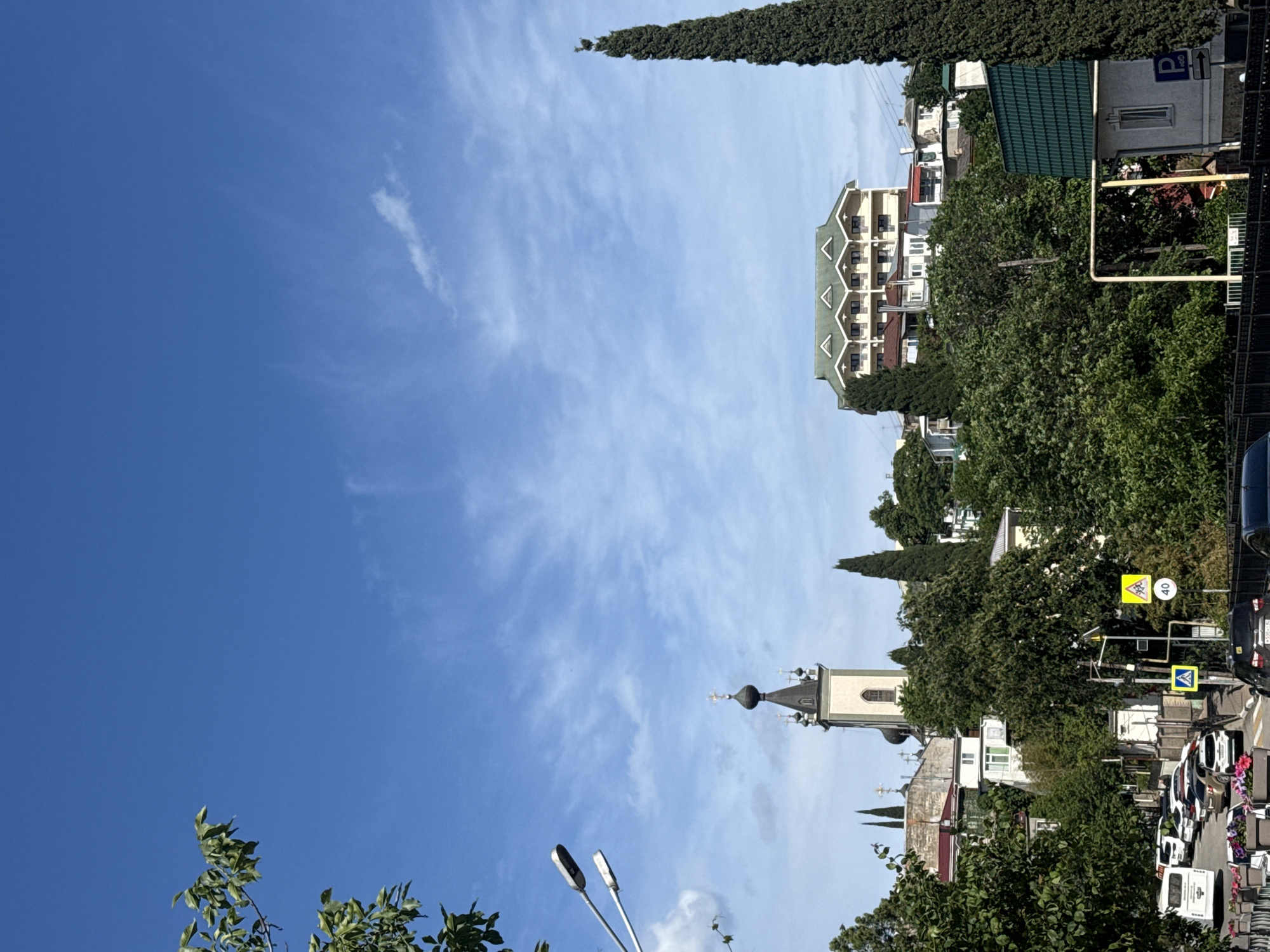 The image captures a cityscape with a blue sky filling the majority of the frame. Wispy clouds are scattered across the sky. Below, a building with a green roof is partially visible. A light-colored structure with multiple floors and green-trimmed balconies is also present. Tall, slender cypress trees punctuate the skyline, adding vertical elements to the scene. A church with a distinctive dome-shaped roof and a cross is visible. Streetlights, trees, and vegetation line the bottom portion of the image, partially obscuring the street and cars. Traffic signs indicate a pedestrian crossing and speed limit. Cars are visible in the lower right corner, suggesting street traffic.