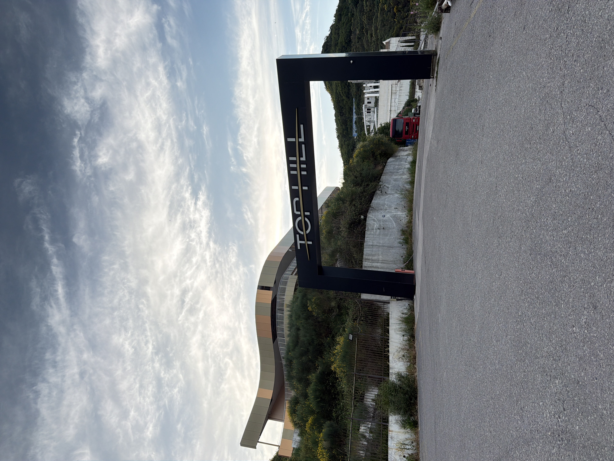 The image depicts the entrance to a property named "TOP HILL" in Budva, Montenegro. A large, black archway with the name "TOP HILL" inscribed in white lettering stands prominently in the foreground. Beyond the archway, a modern, multi-story building with a distinctive curved facade is visible, situated on a hillside covered with lush green vegetation and scattered yellow wildflowers. A red truck is parked on the road leading into the property, with other vehicles and parts of buildings further in the background. The scene is set against a backdrop of a dramatic sky filled with wispy clouds. The overall impression is one of an entrance to a commercial or entertainment venue.