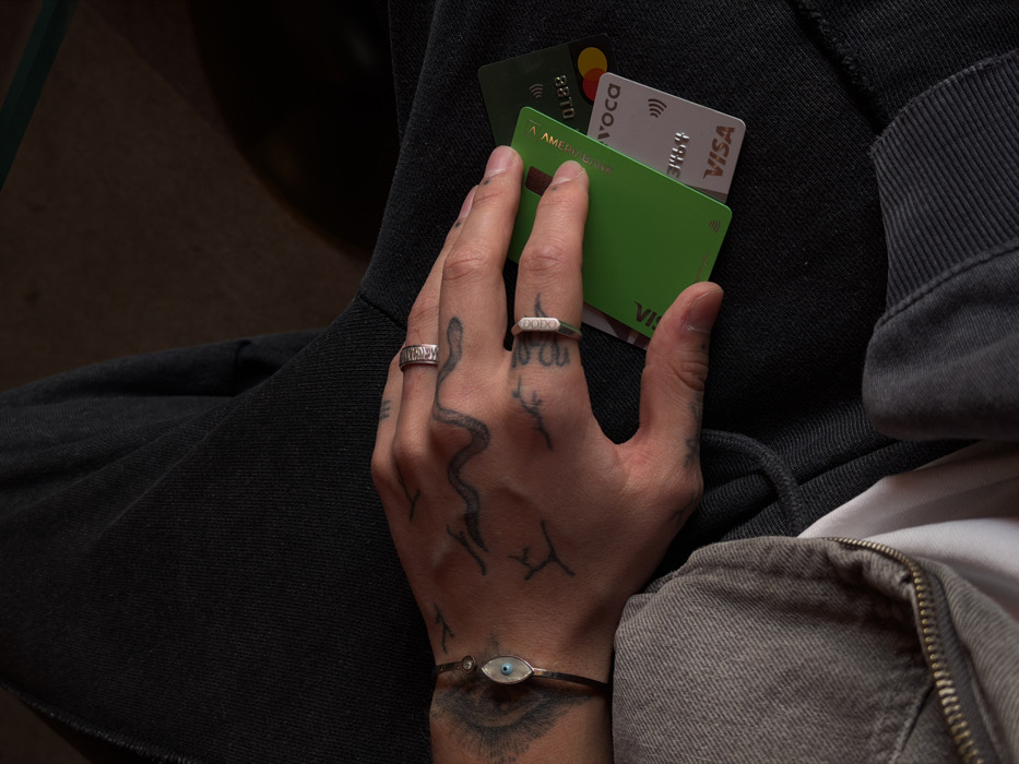 A close-up, overhead shot depicts the left hand and upper thighs of an individual seated indoors. The individual's left hand, displaying multiple tattoos including a snake-like design, holds three payment cards against dark gray or black fabric. A thin metallic bracelet with an oval-shaped evil eye charm is present on the wrist, along with a textured ring on the ring finger and a hexagonal ring on the pinky finger. The payment cards include two bright green cards, one explicitly labeled "AMERIABANK" and "VISA," and another green card with "AMERIABANK" and "VOca" (or "VOCA") visible. A third, partially visible white/silver card features both "VISA" and "MASTERCARD" logos, along with a contactless payment symbol. The individual's left thigh is covered by a lighter gray garment with a visible zipper, revealing a white undershirt beneath. The background is dark and out of focus. The presence of Ameriabank cards indicates the location is in Yerevan, Armenia.