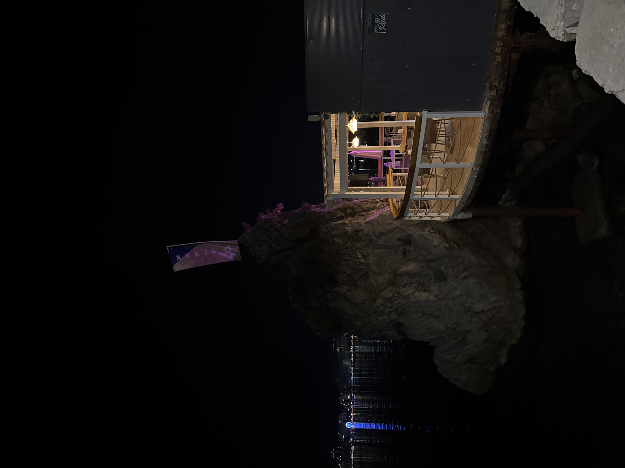 This is a nighttime photograph of a restaurant situated on the water in Budva, Montenegro. To the left, a large rock formation juts out of the dark water, topped with a flag that has purple lettering and the number "40" on it. The city skyline is visible in the distance across the water, with numerous lights and a Ferris wheel illuminated with blue and white lights. The restaurant itself has a covered outdoor seating area with tables and chairs, lit by warm-toned pendant lights. A railing with rope details encloses the seating area. The building facade is dark and corrugated. The overall scene is dark, with artificial lighting creating an ambiance for the dining establishment.