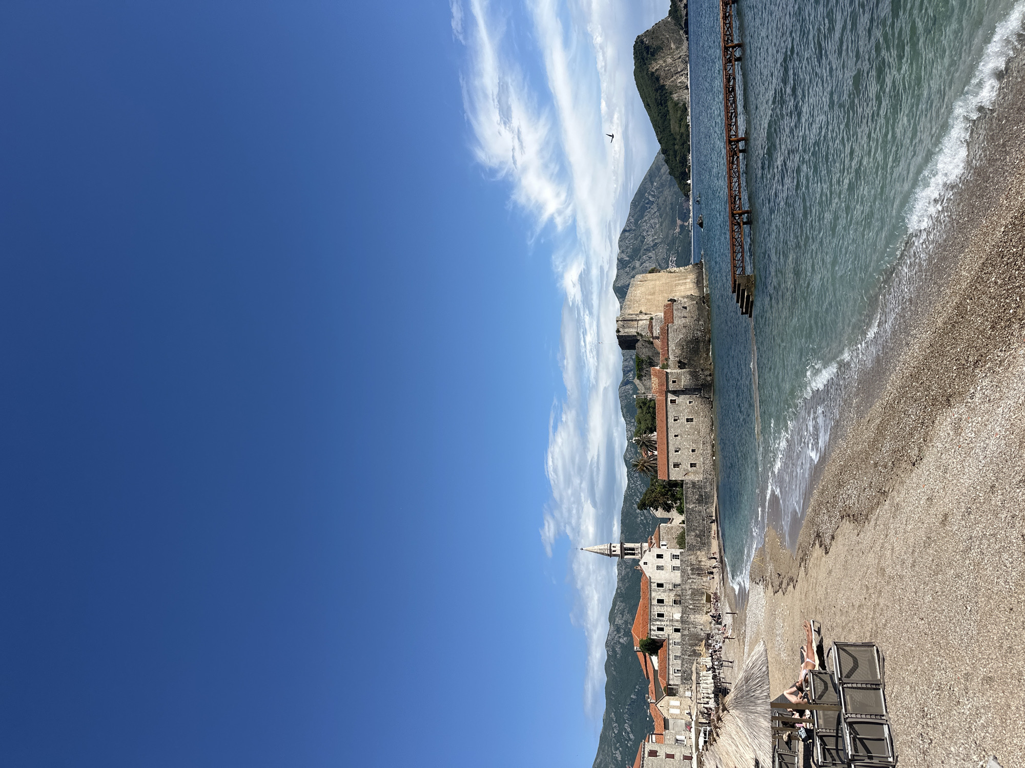 The image depicts the beach area of Budva, Montenegro, during daylight. The sky is predominantly clear blue with some wispy white clouds. To the right, a rocky shoreline meets the turquoise sea with gentle waves breaking on the pebble beach. A rusty metal pier extends into the water. Along the beach, there are sunbathers relaxing on lounge chairs under umbrellas. Ancient stone buildings with red-tiled roofs are clustered together, with a church steeple visible. A mountain range forms the backdrop, partially obscured by clouds. A bird is visible in flight in the sky.