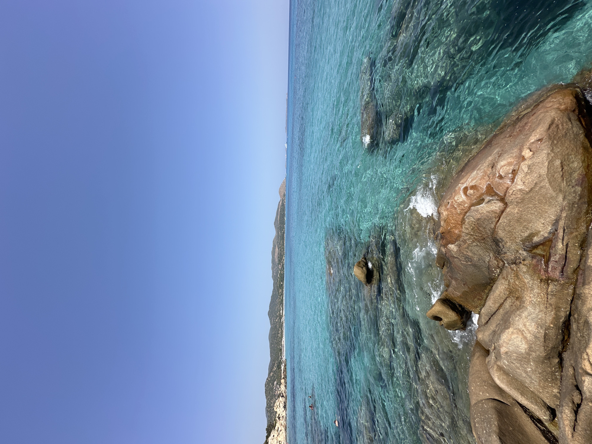 The image depicts a scenic coastal view in the vicinity of Crabonaxa/Villasimius, Italy. In the foreground, large, weathered rocks in shades of brown and orange are partially submerged in the clear, turquoise water. The water is so transparent that the rocky seabed is visible beneath the surface. Small waves gently lap against the shore and the rocks, creating white foam.
Further out, the sea transitions into deeper blue hues. Several people are visible swimming and enjoying the water. In the middle ground, a coastline with sparse vegetation and rocky outcrops stretches across the horizon. Distant hills and mountains form the backdrop, suggesting a Mediterranean landscape. The sky is a clear, bright blue, indicating a sunny day. The overall impression is one of a tranquil and idyllic beach setting.