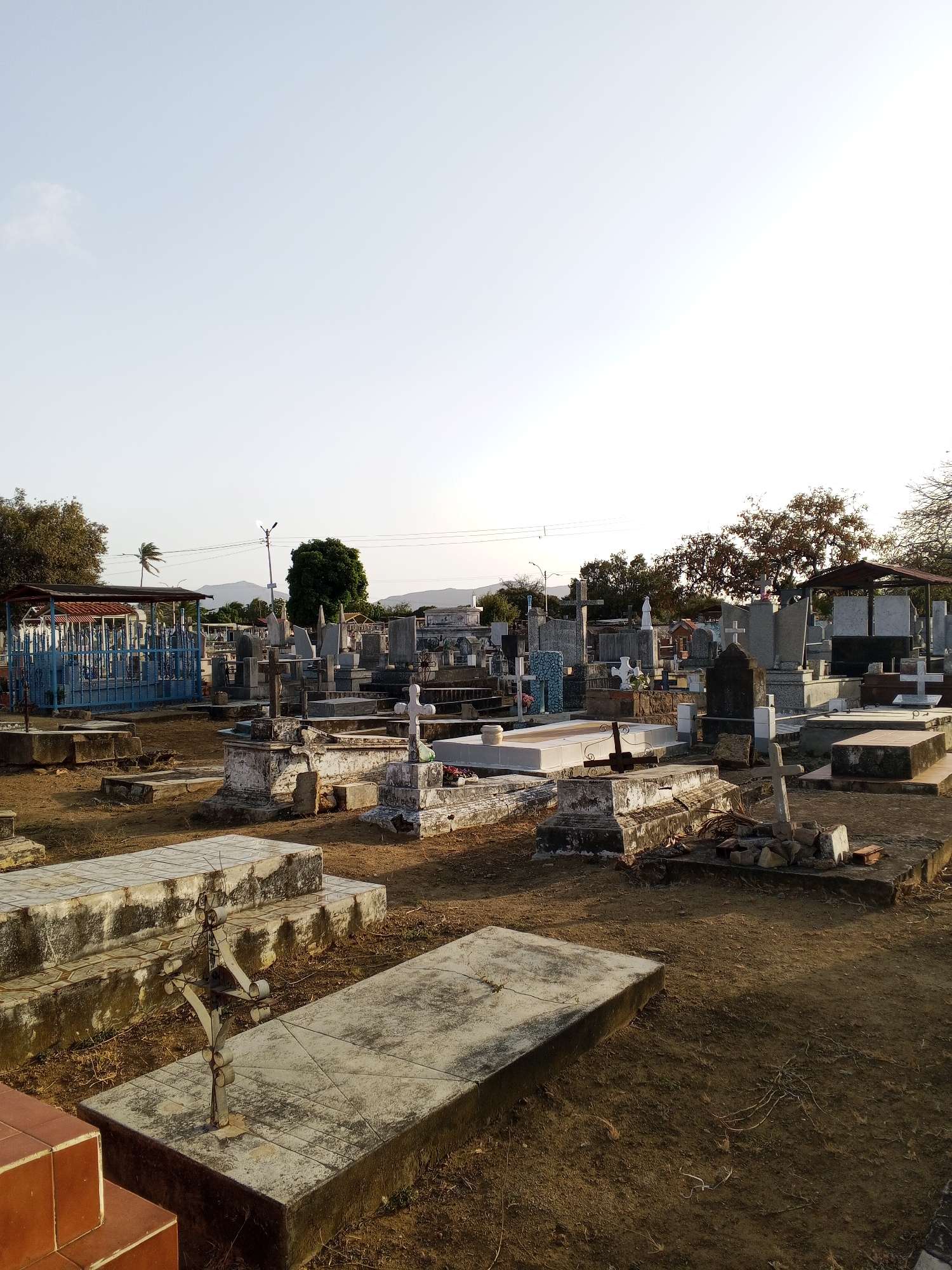 The image shows a cemetery with numerous gravestones and tombs of varying sizes and designs. The ground is mostly dirt and dry grass, with some brick debris near some of the graves. In the background, there are trees and hills, and a clear sky overhead. A blue metal structure covers one of the tomb areas. The location is Unknown, Unknown.