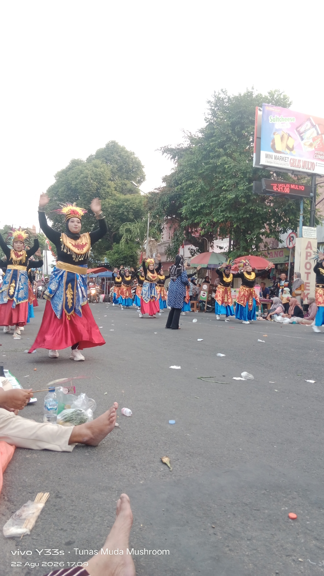 In the city of Genengan, Indonesia, a group of women in traditional attire are participating in a parade. They are dressed in colorful costumes featuring gold, blue, and red fabrics, with elaborate headpieces adorned with feathers. The women are moving in unison, with their arms raised. The parade is taking place on a paved street, with spectators seated on the side. In the background, trees and buildings line the street, and a billboard advertises "Saltcheese." The ground is littered with trash, including plastic bottles and wrappers. The sky is overcast.