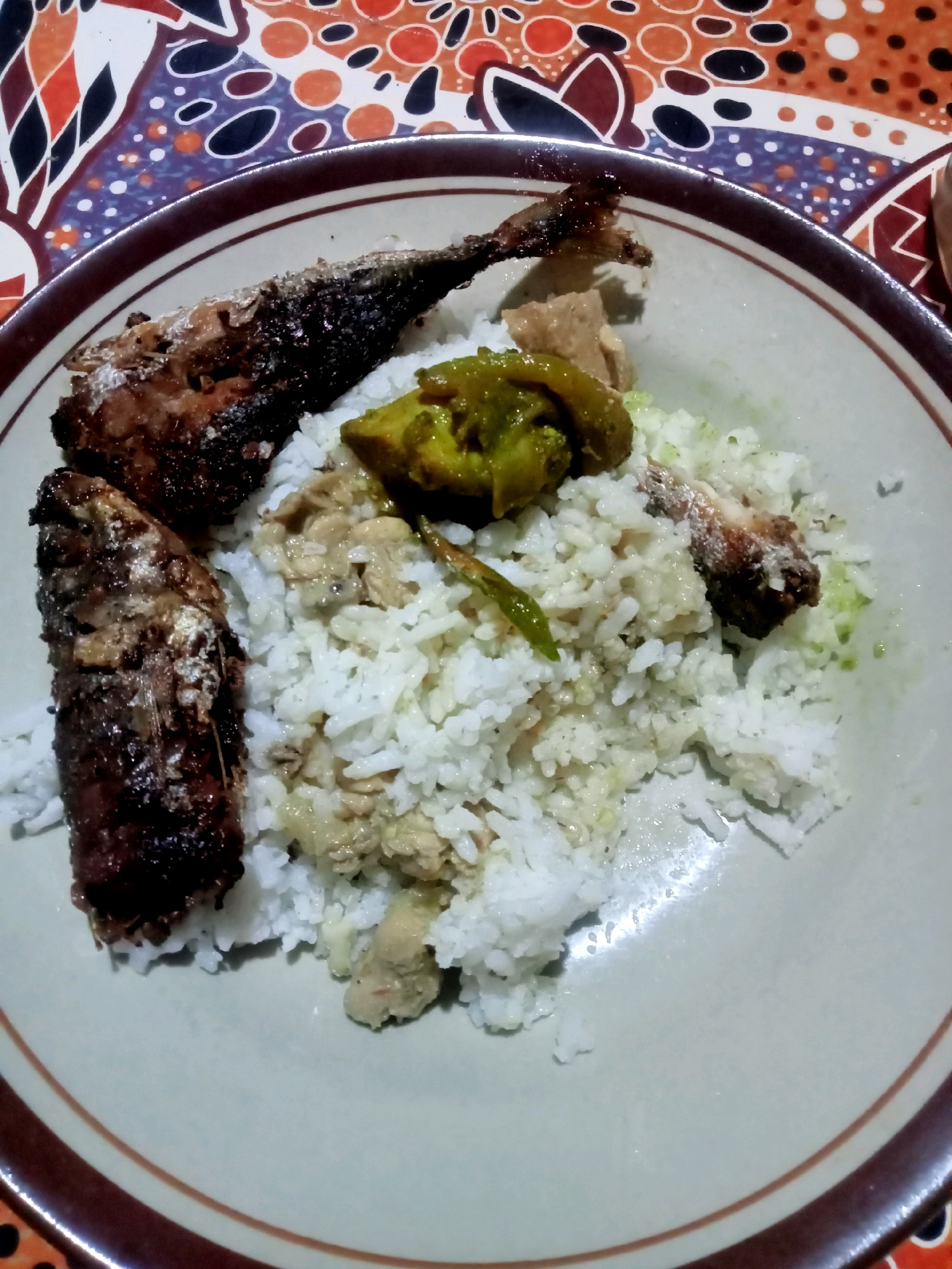 A plate of food sits on a colorful patterned tablecloth. The plate contains white rice, two pieces of fried fish, and a green vegetable dish. The tablecloth has a pattern of orange, red, black, and white shapes. The scene appears to be indoors. This image was captured in Girisubo, Indonesia.