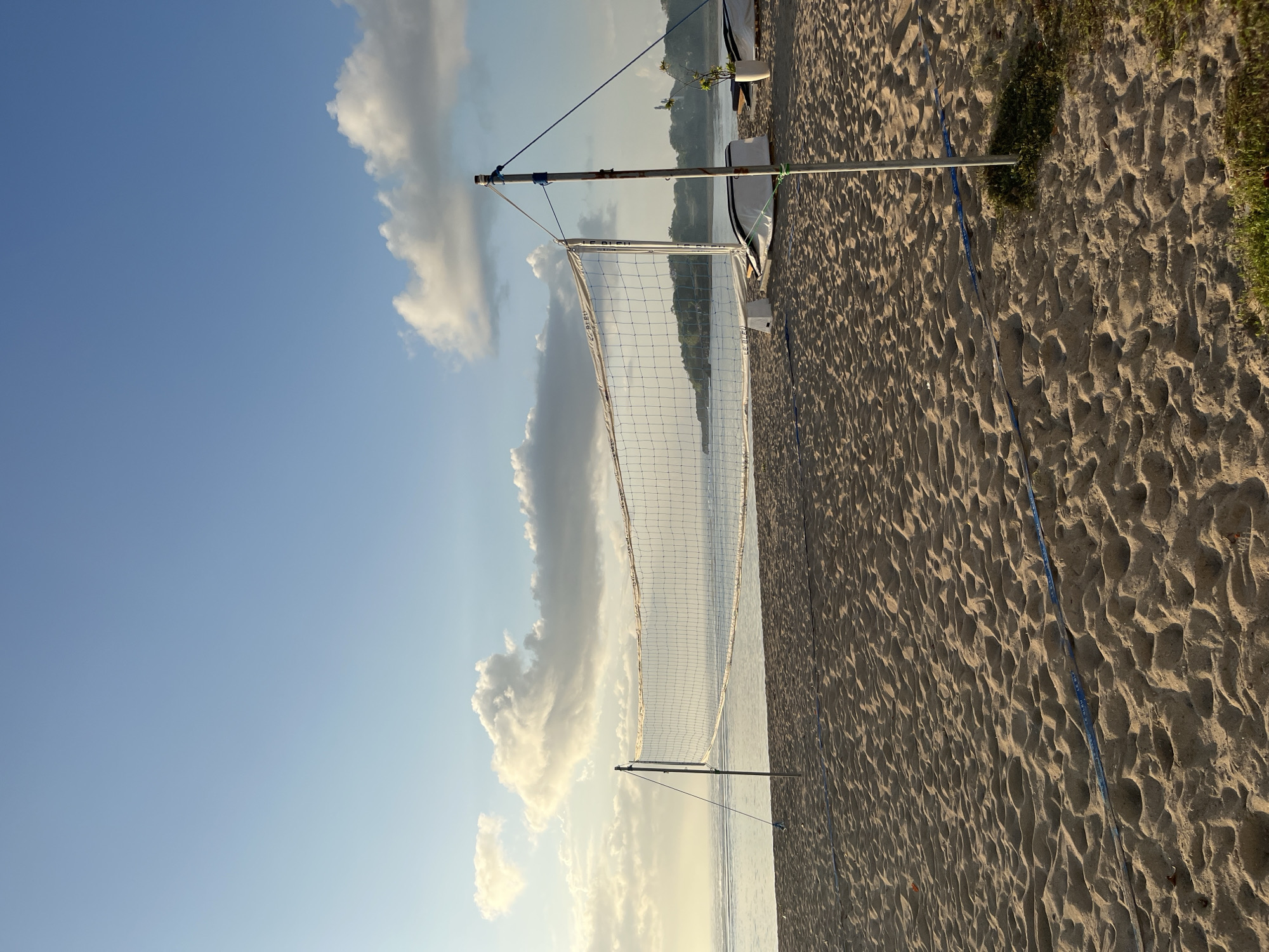 The image depicts a volleyball net set up on a sandy beach in Kuta Selatan, Indonesia. The sand is rippled, indicating the tide or wind. The volleyball net stretches across the frame, supported by two poles. The ocean is visible in the background, with a calm surface and a distant shoreline lined with greenery. The sky is a clear blue with scattered white clouds, suggesting it is daytime. There are no people or active events captured in the scene. Some white objects, possibly parts of beach chairs or equipment, are present near the net on the right side. A white vase with a plant is also visible. The overall atmosphere is serene and tranquil.