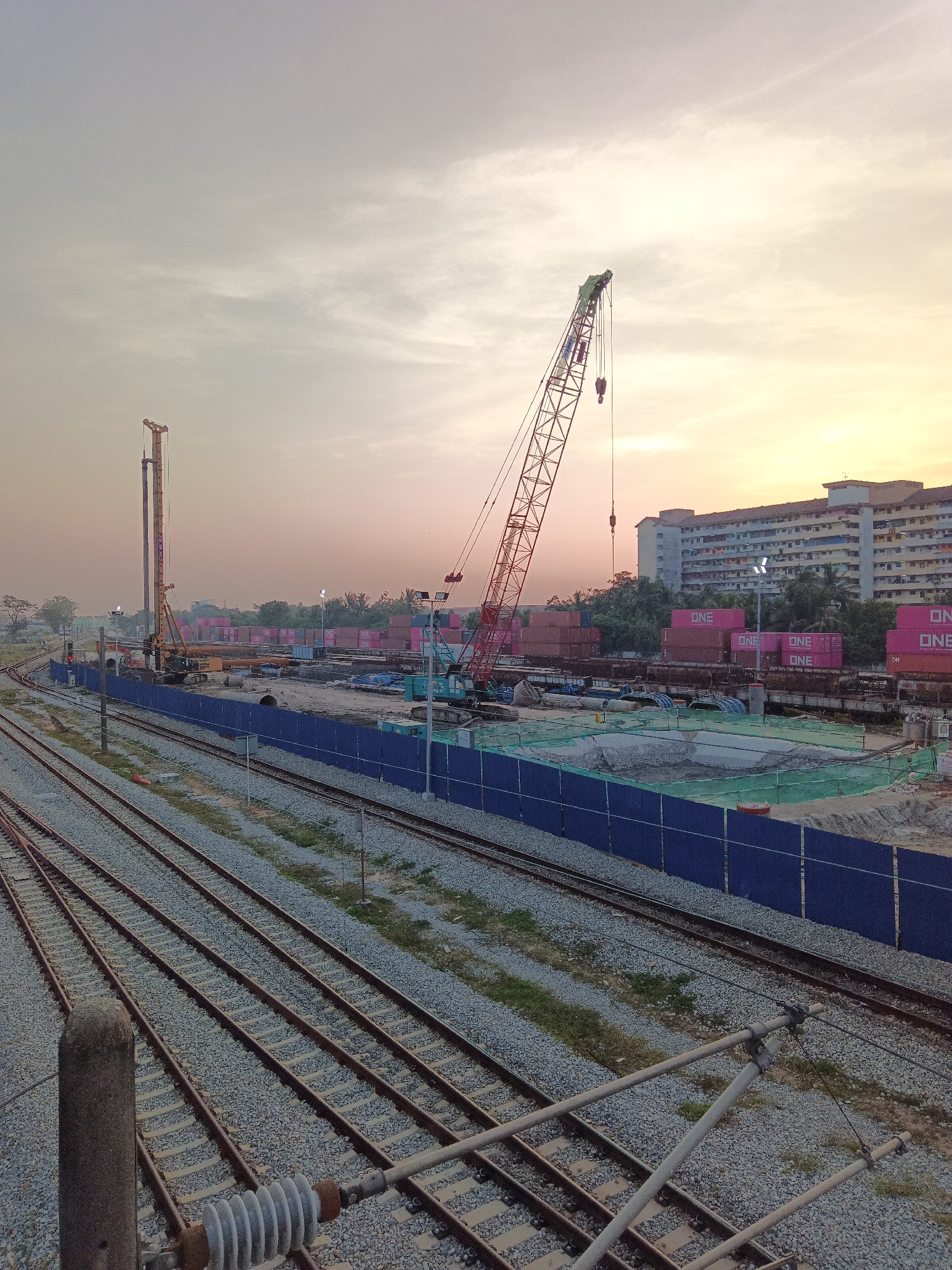 This image captures a construction site at dusk in Klang City, Malaysia, adjacent to a railway line. The scene is dominated by a large red construction crane positioned in the middle ground, with its boom extended upwards. To the left, a yellow piling rig stands tall, also a part of the ongoing construction.
In the background, several rows of shipping containers, many marked with "ONE," are stacked. Behind the containers, a multi-story residential building with many windows is visible, suggesting the proximity of a residential area to the industrial or construction zone. The sky is a soft gradient of orange and grey, indicating the transition from day to night.
The foreground features multiple sets of railway tracks, with ballast and vegetation visible between them. Electrical poles and wires for the railway system are also present. A blue construction fence delineates the boundary of the construction site. The overall atmosphere is one of industrial activity and urban development during the evening.