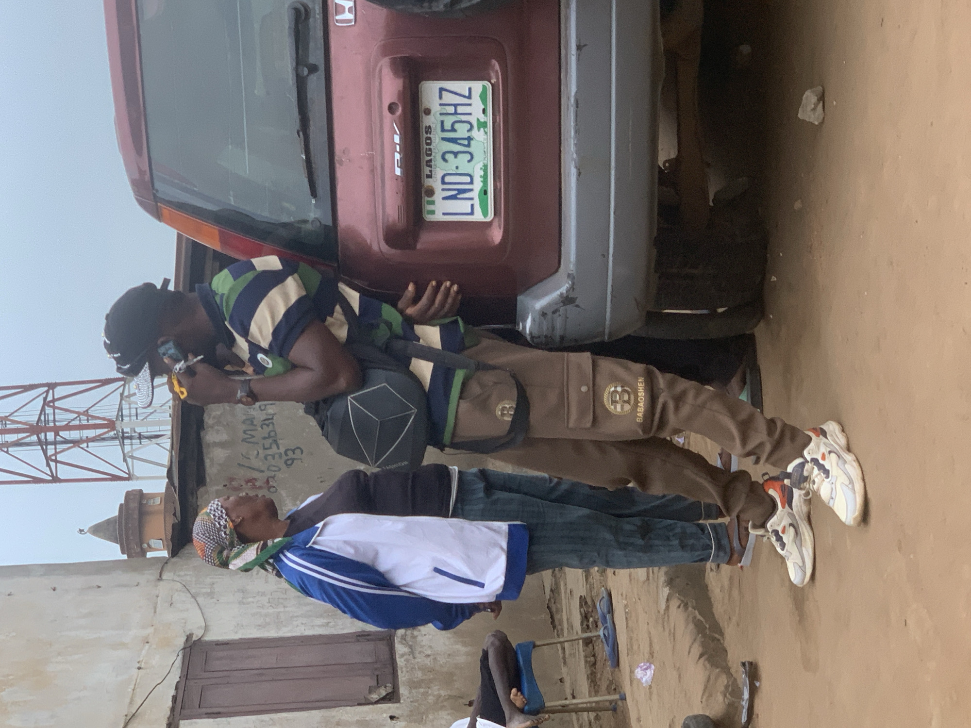 The image depicts an outdoor scene with several individuals and a vehicle. In the foreground, a man wearing a striped shirt, brown cargo pants with the inscription "BABAOSHEN", and athletic shoes is standing next to a dark red SUV. He is holding a mobile phone to his ear. Behind him, to the left, a woman with a headscarf, a white and blue jacket over a dark shirt, and patterned trousers is standing and looking towards the man. To the left of the woman, a person is seated on a blue chair. The SUV has a license plate that reads "LAGOS LND-345HZ". In the background, a building with a window and a distinct architectural element resembling a mosque dome with a crescent moon is visible, along with a large red and white communication tower. The ground appears to be unpaved, with loose dirt and some scattered debris. The overall environment suggests a casual, possibly informal, setting within a Nigerian city, with the presence of the "LAGOS" license plate indicating a connection to that state. Given the context provided that the location is Shagamu, Nigeria, the scene can be interpreted as a street-level view in that city, potentially near residential or commercial areas, with characteristic urban infrastructure and vehicle presence.