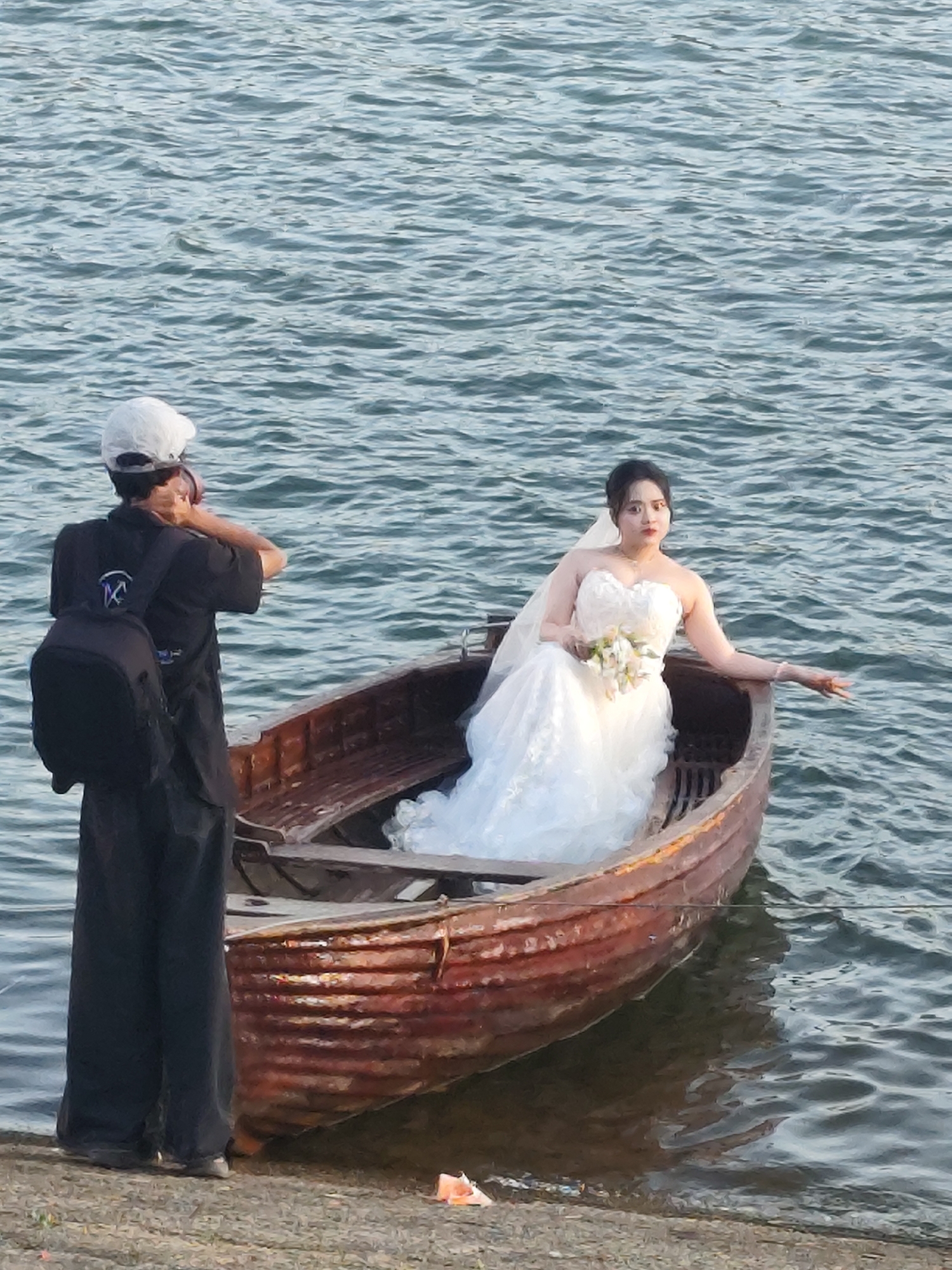 In a waterfront scene, a person wearing a white cap, black shirt, and dark pants stands facing away from the camera, holding a camera to their face, as if taking a photo. They are wearing a black backpack. The person is positioned on the left side of the frame, next to a wooden boat filled with water. Inside the boat, a woman in a white wedding dress with a veil and holding a bouquet of flowers is seated, looking to her right. Her left arm is extended. The boat is near the shore, with the water reflecting the light. The environment suggests an outdoor location with a body of water.
