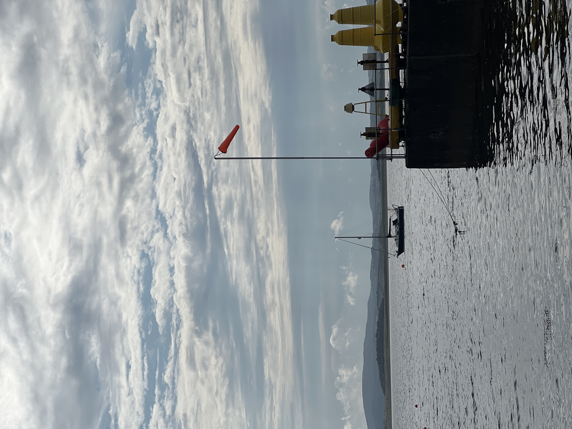The image depicts a body of water with a cloudy sky. In the foreground, a dark, rectangular structure, likely a pier or dock, occupies the right side of the frame. On top of this structure are several yellow, conical buoys, a red conical object, and a metal pole with an orange windsock. In the middle ground, a sailboat with dark hulls and white sails is visible on the water. Further in the distance, rolling hills and a shoreline are present. The water is choppy, and there are a few small red buoys scattered on its surface. The overall scene suggests a coastal or harbor area, possibly in or around Terhillion, Ireland, given the context. There are no people or discernible interactions visible in the image.