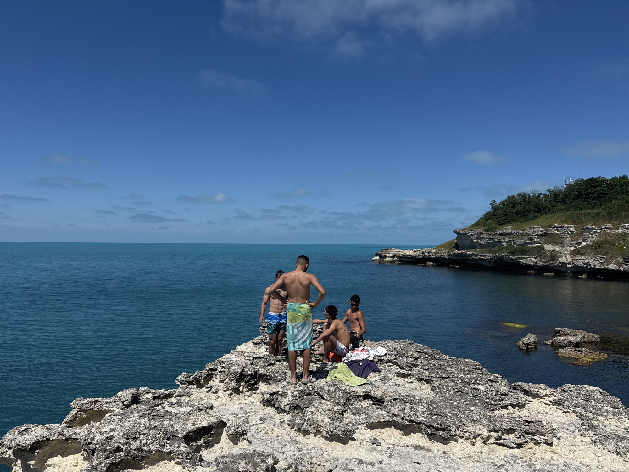 The image depicts a group of four young males on a rocky outcrop overlooking the sea. The location is identified as a cliffside area near Tsarevo, Bulgaria. The sky is clear blue with scattered clouds. The sea is a deep turquoise, calm and expansive. The rocky terrain is uneven and appears to be composed of light-colored stone, characteristic of coastal erosion.
One individual, standing with his back to the camera, wears a light blue and white striped towel around his waist. To his left, another young man in patterned swim trunks stands. To the right of the towel-clad figure, a third individual is seated on the rock, wearing dark shorts. A fourth young man, also in dark shorts, is standing further to the right, facing away from the viewer. Scattered on the rocks are towels and possibly swimwear. The general atmosphere suggests a recreational outing, likely on a warm day. The background shows a rugged coastline with lush green vegetation on the higher ground, and the distant horizon where the sky meets the sea.