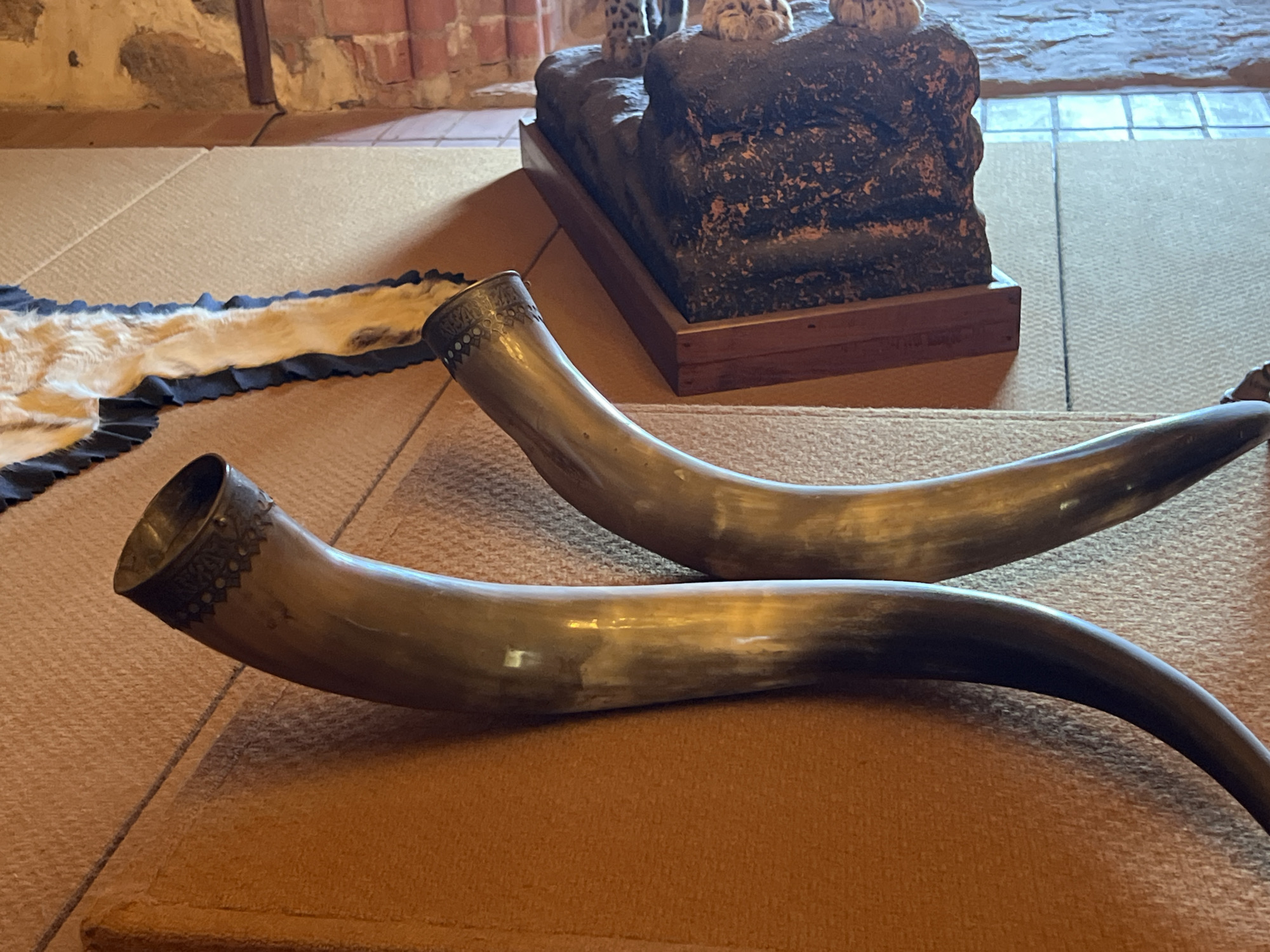 The image shows two ornate drinking horns, possibly made from animal horns, placed on a textured brown mat. One horn is positioned in the foreground, curving to the right, while the other is further back and to the left, also curving to the right. Both horns have decorative metal bands around their openings, featuring intricate patterns. Behind the horns, a piece of fur with dark edging is laid out on a similar mat. In the background, a stone wall with visible brickwork can be seen, and a dark, rough-textured object on a wooden stand, possibly a decorative sculpture, is present. The scene appears to be indoors, with the lighting suggesting it might be a dimly lit room or a location with natural light filtering in. The overall impression is one of historical or cultural artifacts displayed in a setting that might be a museum, a historic building, or a themed environment. Considering the mention of Trakai, Lithuania, this could be a display within Trakai Castle or a related historical site, showcasing artifacts reminiscent of medieval or ancient traditions.
