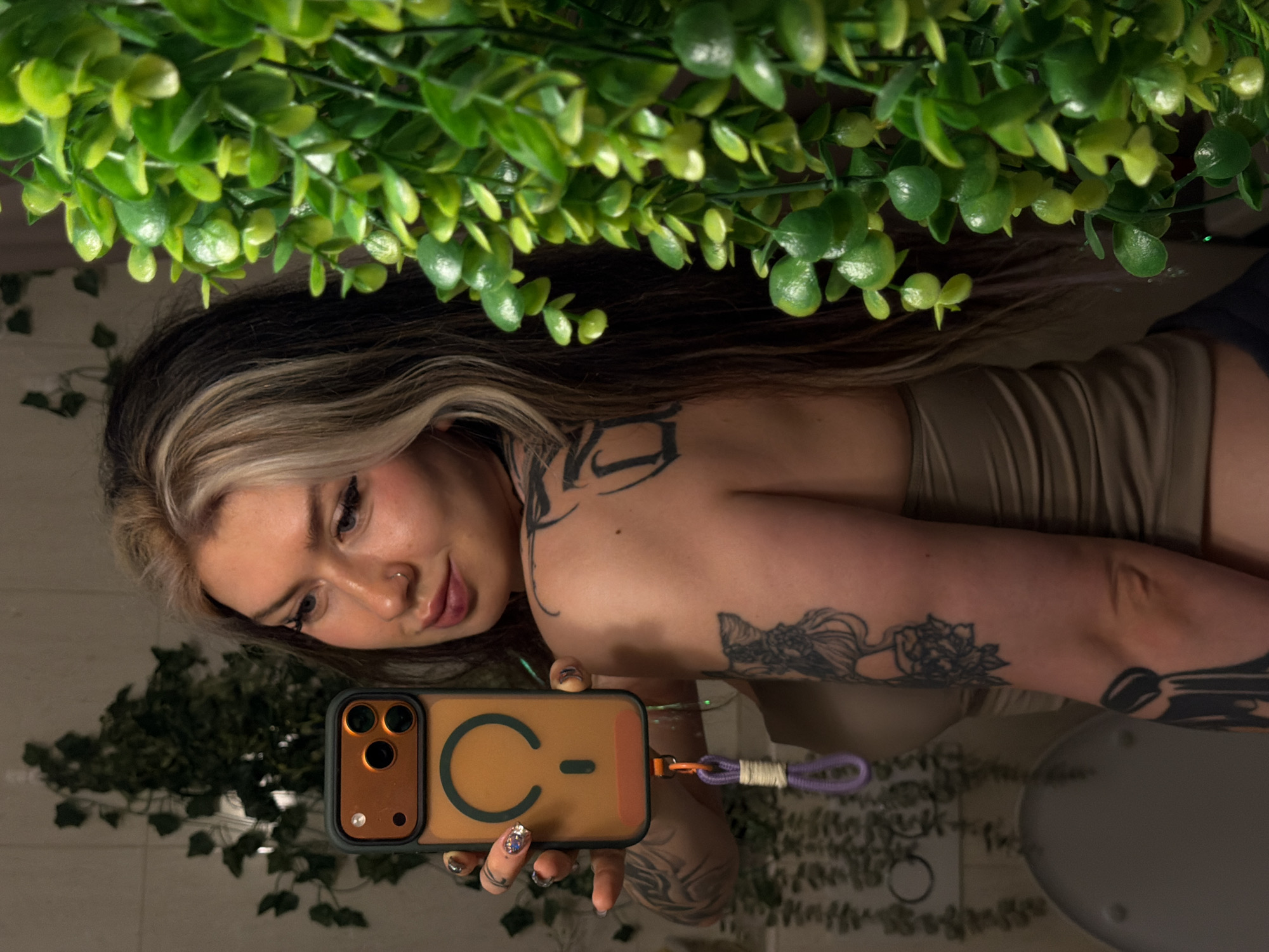 A woman with blonde-streaked dark hair and tattoos is taking a selfie in what appears to be a bathroom. She is holding an iPhone with an orange case and a purple wrist strap. The background features artificial green plants on the walls and ceiling, and a toilet seat is visible on the right. The location appears to be a private residence in an unspecified location within Budva, Montenegro.
