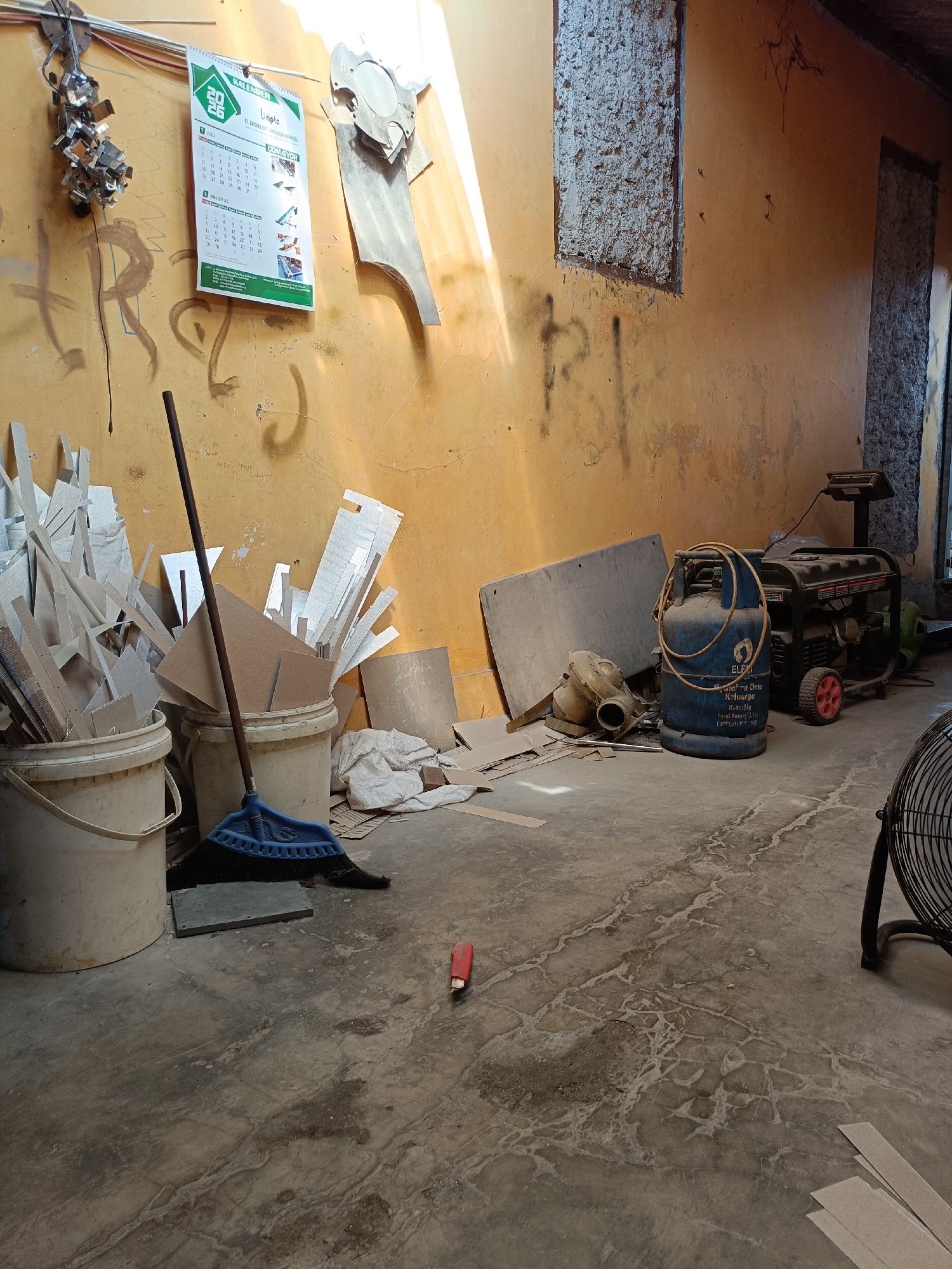 The image depicts an interior space, possibly a workshop or storage area, with a light-colored concrete floor and an orange-yellow painted wall. The wall features graffiti in dark spray paint. Sunlight streams in from an unseen source, casting strong shadows and illuminating parts of the room.
Prominent objects in the scene include:
* **A generator:** A black and red portable generator is positioned on the floor, with its exhaust pipe visible.
* **A gas cylinder:** A blue propane gas cylinder with a coiled hose rests near the generator.
* **Buckets and debris:** Two white plastic buckets are filled with various pieces of light-colored material, likely wood or cardboard scraps. A blue broom with a dark handle leans against one of the buckets.
* **A calendar:** A white and green calendar for the year 2026 is hanging on the wall.
* **Metal objects:** A cluster of shiny metal pieces, possibly fasteners or small machine parts, is attached to the wall near the top left.
* **A fan:** The back of a black oscillating fan is visible on the right side of the image.
* **A utility knife:** A red utility knife lies on the floor in the center of the image.
* **Various scraps:** Numerous pieces of cardboard and other flat materials are scattered on the floor and leaning against the wall.
The environment appears somewhat disorganized, suggesting a work-in-progress or a utilitarian space. The lighting and the presence of tools and materials indicate an active or recently used area. The location of Telagamurni, Indonesia, is mentioned, but no specific identifying landmarks within the image confirm this.