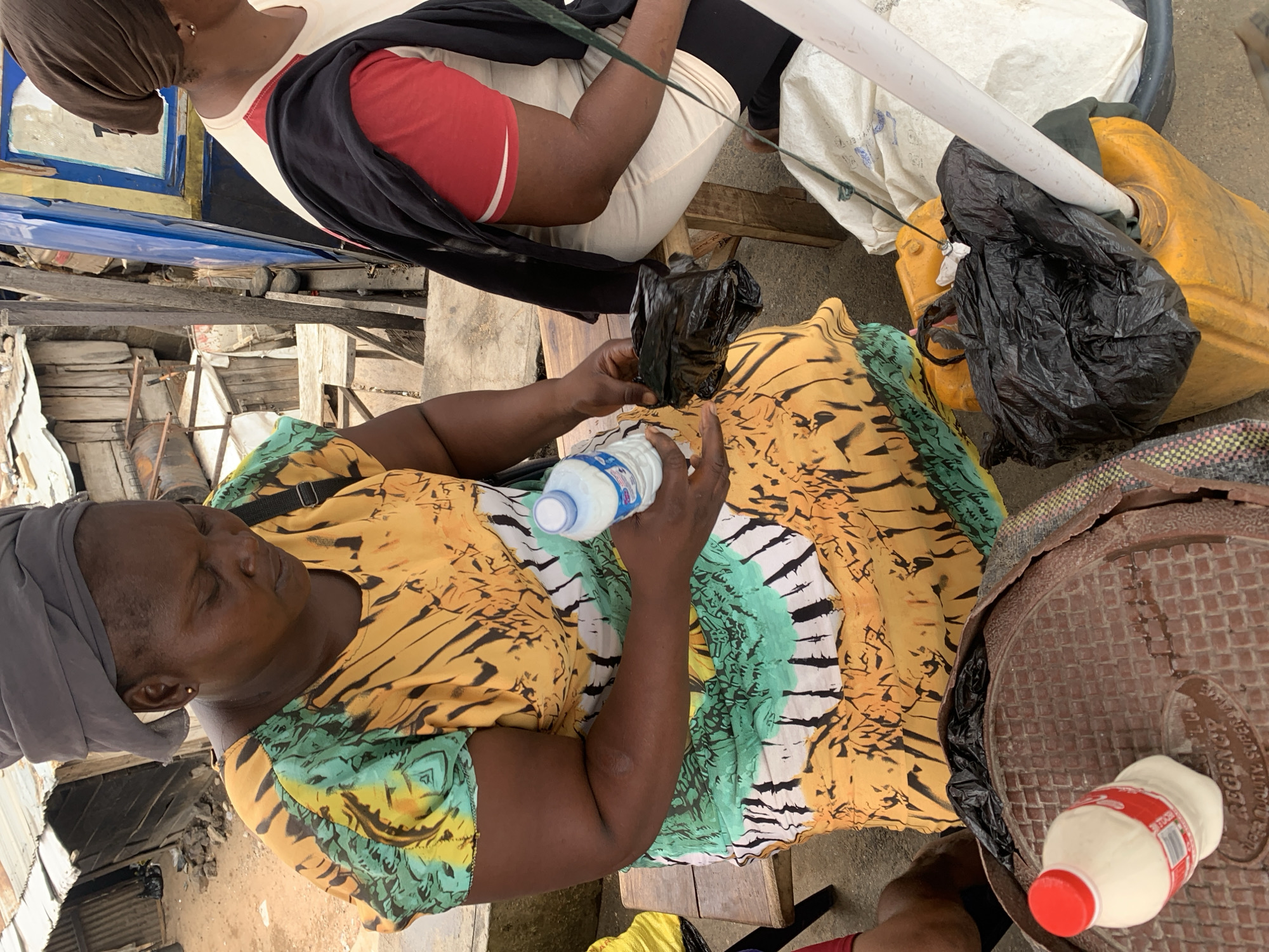A high-angle shot depicts an outdoor scene in Lokoja, Nigeria. An adult woman with a gray headwrap sits on a wooden stool, holding a water bottle and a black plastic bag. She wears a yellow and green patterned dress. Next to her, another person sits, wearing a beige shirt with a red sleeve and a black top draped over it. Beside them are a yellow jerrycan, a white plastic pipe, and a brown circular object, with a white bottle with a red cap placed on top. The surroundings consist of corrugated metal and wood, suggesting a market or informal settlement.