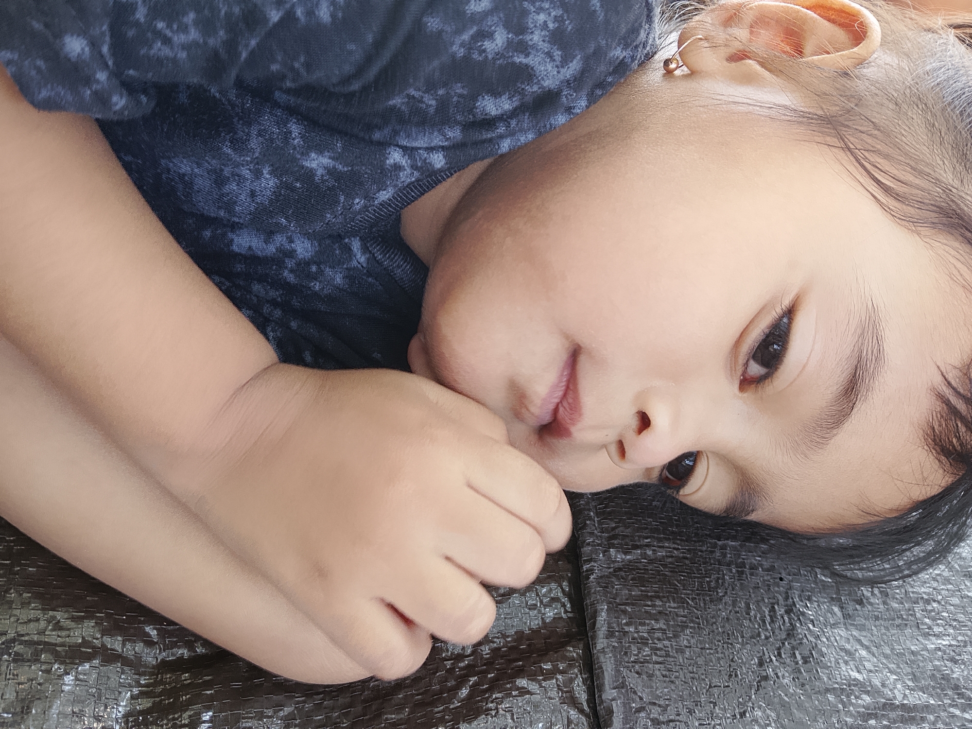 The image shows a close-up view of a young child lying down, with their head resting on their hands. The child is wearing a dark, patterned shirt. Their face is turned to the side, and their eyes are looking away from the camera. The background appears to be a dark, textured surface. There is no discernible location within the city of Ngadrin, Indonesia.
