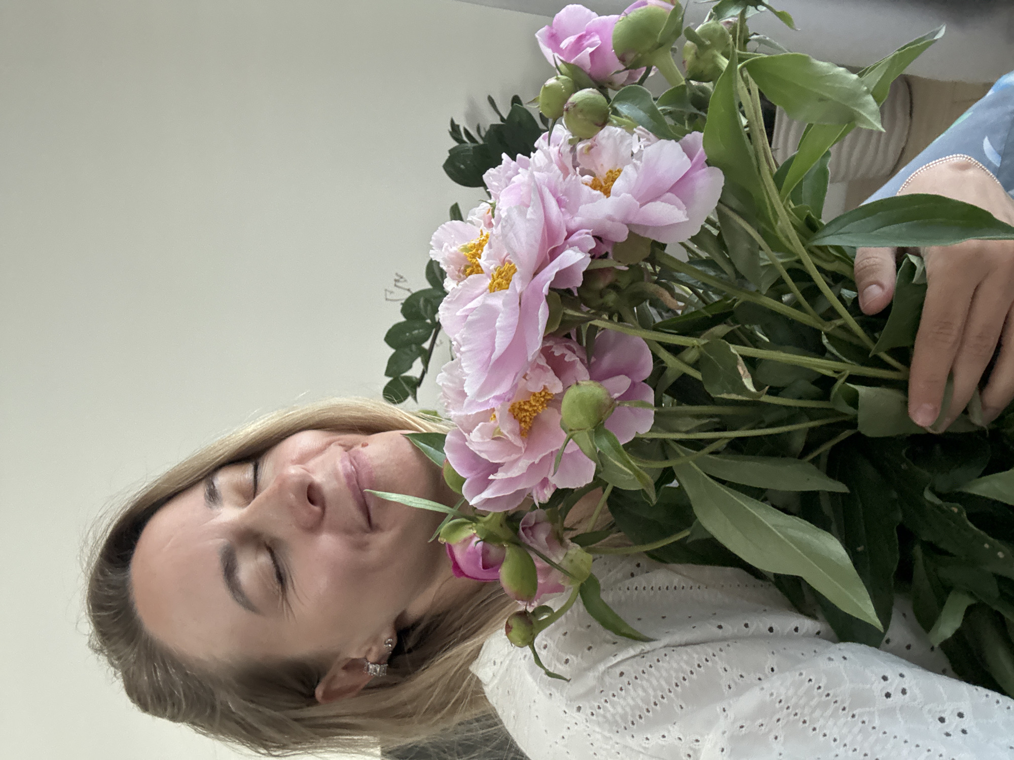 In this media file, a woman is captured in a close-up shot, holding a bouquet of light pink peonies. Her eyes are closed, and she has a serene expression, suggesting a moment of peace or enjoyment. She is wearing a white, eyelet-patterned top. The background is a plain, light-colored wall, with a hint of a blue patterned fabric visible in the upper right corner, possibly part of her clothing or surroundings. The lighting appears to be natural, suggesting daytime. The overall mood is tranquil and intimate, focusing on the woman and the beauty of the flowers.