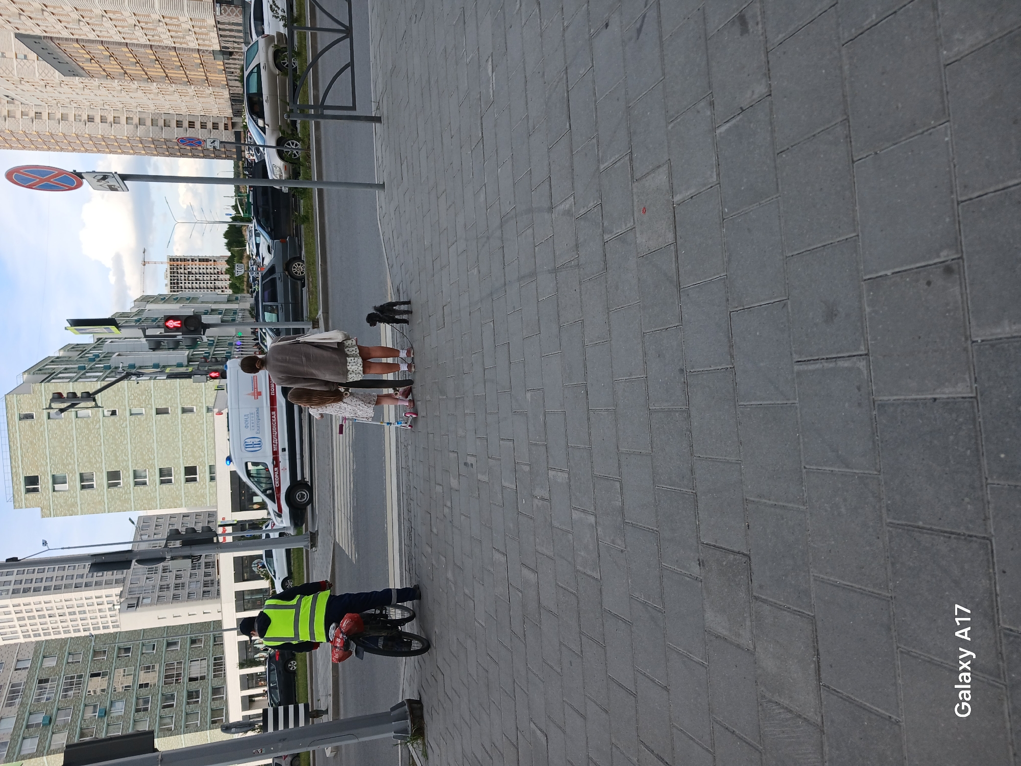 The image shows a street scene with several people. A woman and a girl are standing on the sidewalk with a small dog. A cyclist wearing a yellow vest is positioned at the edge of the sidewalk near a crosswalk. An ambulance is parked in the background. The setting appears to be in an urban area of Yekaterinburg, Russia, as suggested by the ambulance markings. Traffic lights can be seen in the scene, indicating a street intersection. The overall impression is of a casual moment in a city environment.