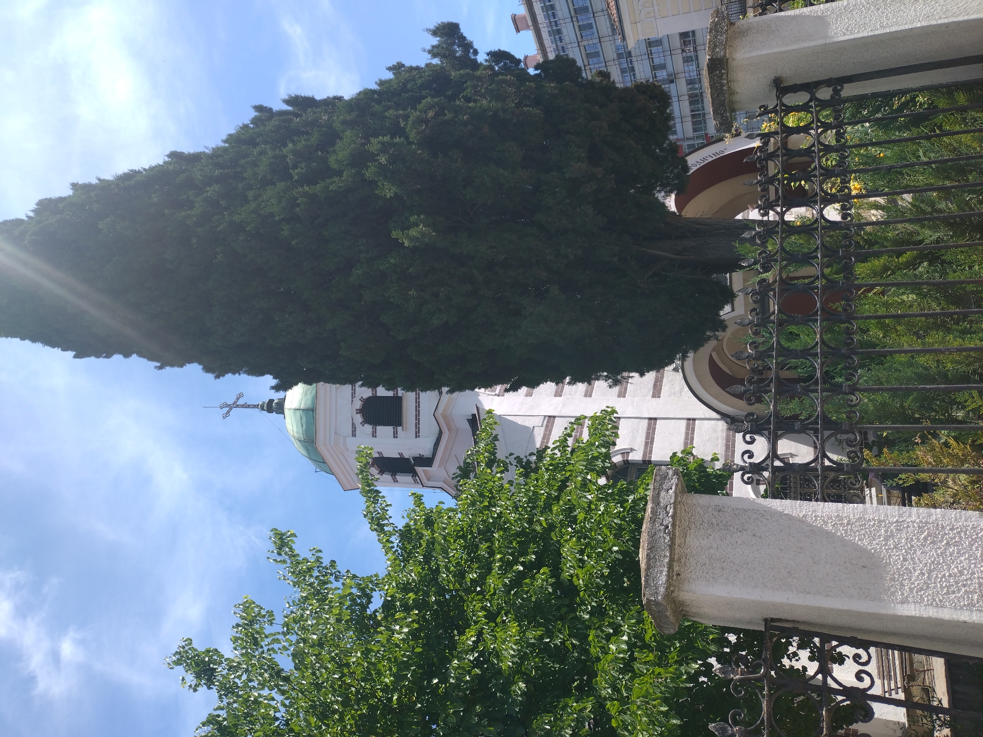 The image depicts a view of the Sveti Nikola (Saint Nicholas) Church in Stara Zagora, Bulgaria, from a lower angle, looking upwards towards the church's dome and cross. The church is partially obscured by trees and a wrought iron fence. The church's facade is white with dark vertical stripes and a brown roof with a green dome and a cross at the top. A tall, dark green tree dominates the top of the image, obscuring the top of the church building. In the background, a multi-story building can be seen. A concrete wall and wrought iron fence are partially in the foreground, with green foliage visible behind the fence. The sky is blue with wispy clouds.