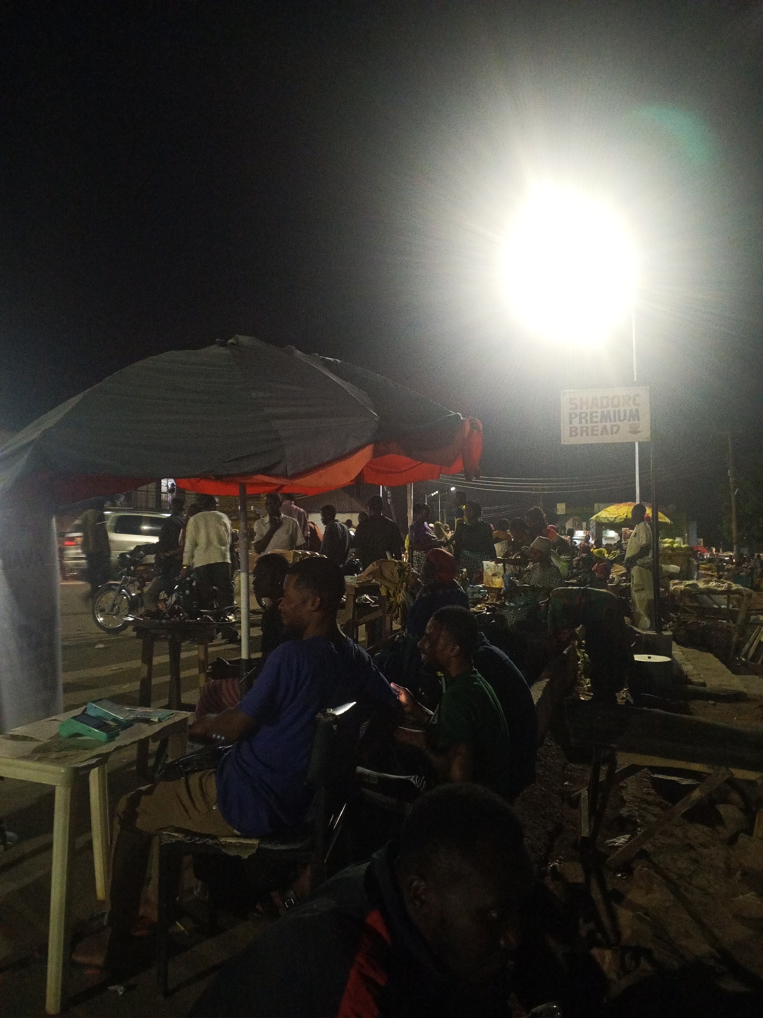 The image captures an outdoor scene at night in what appears to be a busy public space in Gwong, Nigeria. A bright light source dominates the upper portion of the image. In the foreground, people are seated at tables under large umbrellas. Some appear to be engaged in conversation. A sign reading "Shadowe Premium Bread" is visible in the background. A motorcycle and a white car are seen on the road. Numerous individuals are milling about in the background, amidst what seems to be a market area with various stalls and objects.