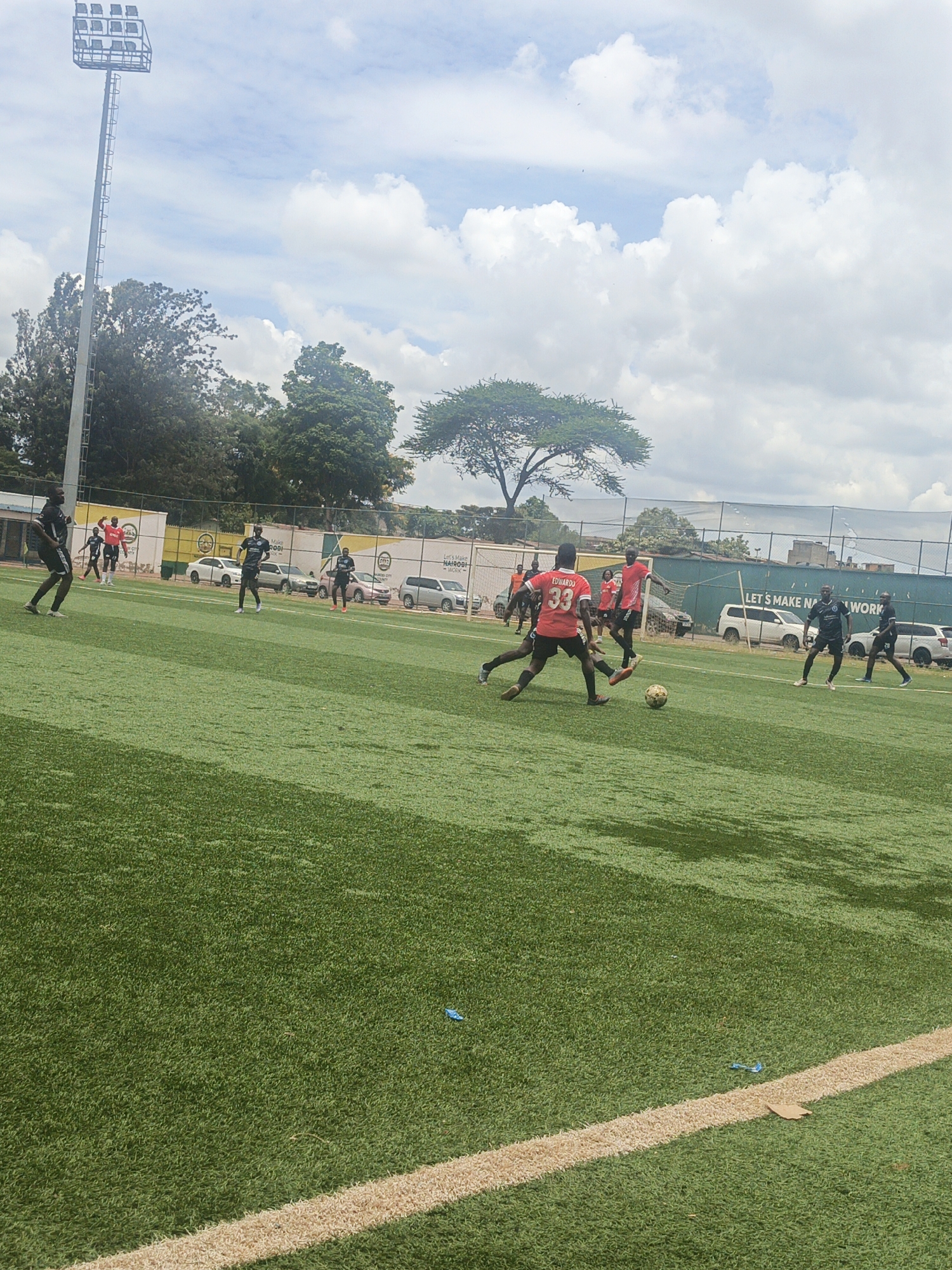 The image depicts a soccer game in progress on a green artificial turf field. Several players are visible, some wearing red jerseys and others in black. One player in a red jersey labeled "33" is kicking the soccer ball. In the background, there is a white fence with signage including the words "LET'S MAKE NAIROBI WORK" indicating this is likely in Nairobi, Kenya. Cars are parked along the fence. Trees and a stadium light tower are also visible in the background under a cloudy sky.