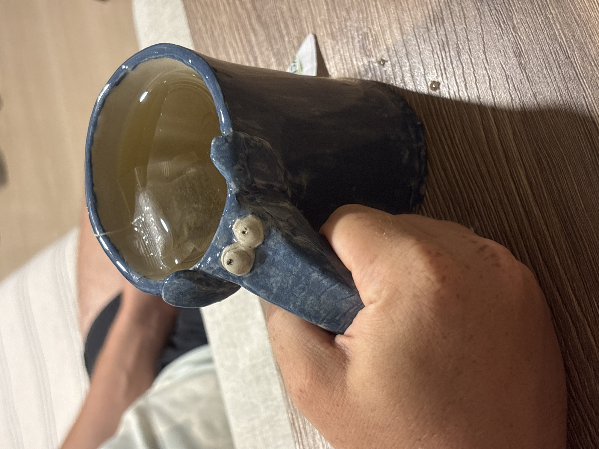 A person is holding a blue ceramic mug filled with tea and a tea bag. The mug has a whimsical design featuring raised "eyes" near the handle. The background includes a wooden surface and part of a white upholstered object, possibly a sofa or chair. There is no indication of the location being Choeng Thale, Thailand.