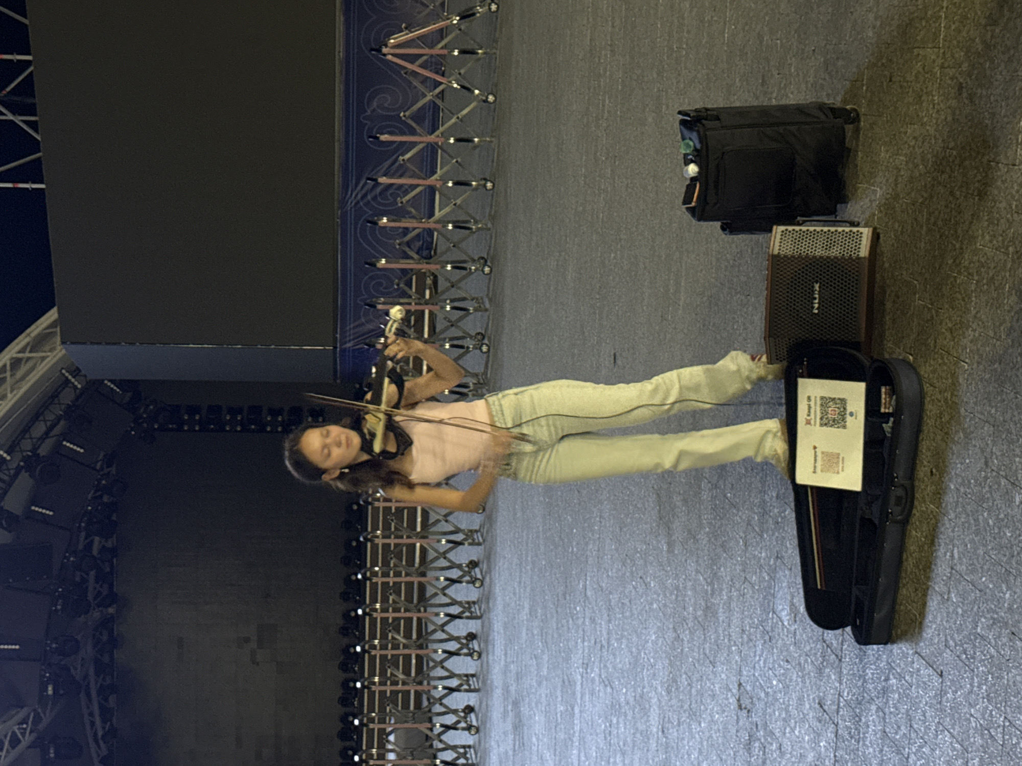 A young woman stands in an open paved area, playing a violin. She is wearing a light pink tank top and light-colored jeans. Her violin is held under her chin, and she is drawing the bow across the strings. A black violin case is open on the ground in front of her, displaying a QR code. To her right, there is a small amplifier labeled "NUX" and a black rolling bag with several small objects on top. In the background, there is a large, dark screen, possibly part of an outdoor stage setup, and rows of portable barriers. The scene appears to be outdoors at night, with artificial lighting evident from above. The overall impression is of a street performer in an urban setting, likely in Pavlodar, Kazakhstan, given the information provided.
