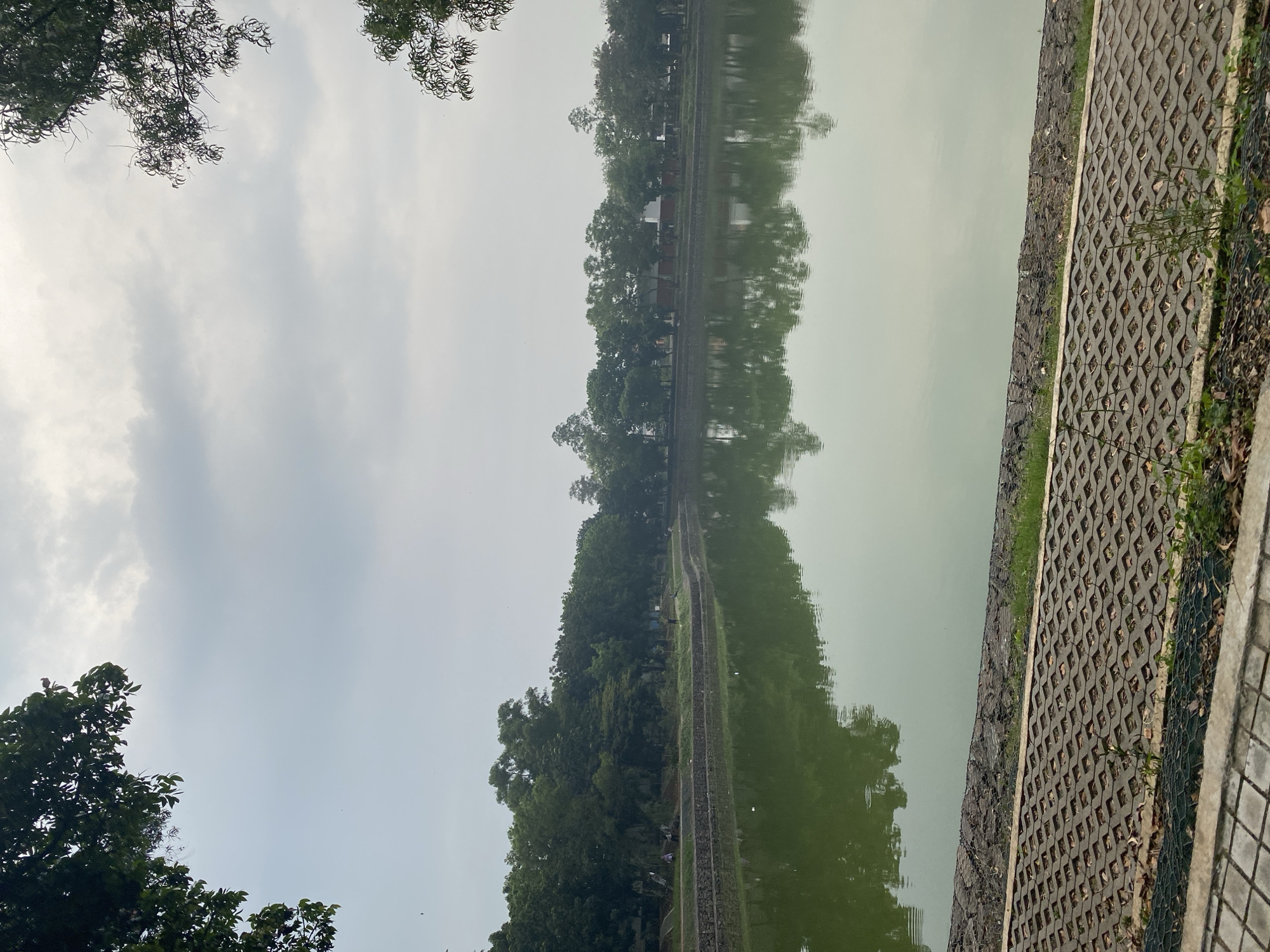The image depicts a scenic view of a calm lake on an overcast day. Lush green trees line the opposite bank of the lake, with some buildings partially visible behind them. The trees also frame the upper corners of the image, suggesting a park-like setting. The water reflects the sky and the surrounding trees, creating a symmetrical and tranquil scene. In the foreground, a textured concrete pathway runs along the edge of the lake, separated from the water by a low, rocky embankment. The overall atmosphere is peaceful and serene, characteristic of a park or recreational area. This scene is likely located in Taman Mini Indonesia Indah (TMII) in East Jakarta, Indonesia, a popular tourist destination known for its themed areas and abundant green spaces, including various lakes and gardens.