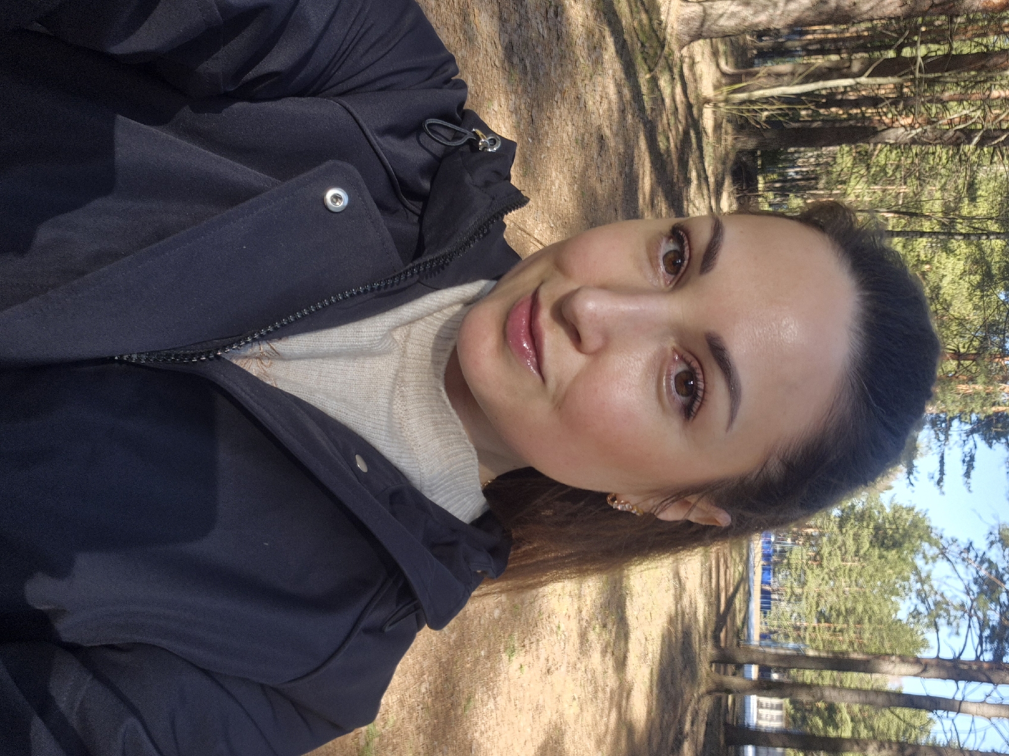 The image is a selfie of a woman, oriented vertically (originally rotated 90 degrees counter-clockwise). The woman is positioned in the foreground, looking directly at the camera with a gentle, faint smile. She has dark hair pulled back, brown eyes, and is wearing natural-looking makeup, including lip gloss and eye makeup.
She is dressed in a dark-colored jacket, likely navy or black, which features a zipper closure and silver-toned snap buttons visible on the placket. Beneath the jacket, a light-colored, possibly beige or off-white, knitted sweater is partially visible. Small, decorative earrings, appearing gold-toned or beaded, are visible in her ears.
The background depicts an outdoor, natural setting, strongly resembling a forest or wooded area. Numerous tree trunks, possibly pine trees, are visible, casting distinct shadows on the ground, indicating direct sunlight. The ground is covered with what appears to be dry leaves, pine needles, or dirt. Further back, green foliage of trees is discernible under a clear, bright sky. The overall impression is a sunny day in a natural, serene environment. There is no visible text in the image.