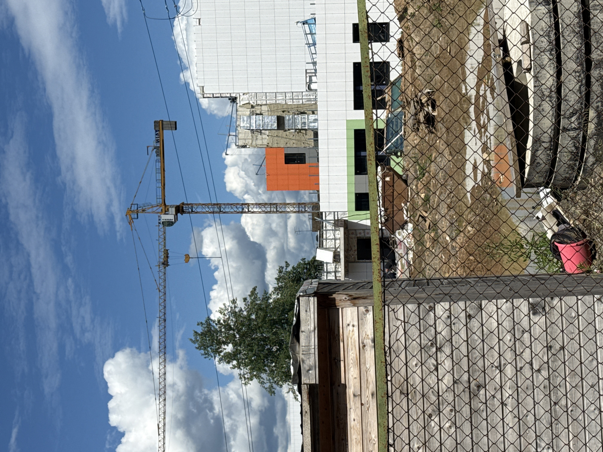This is an outdoor, eye-level, medium shot photograph taken on a cloudy day. In the foreground, a weathered wooden structure with a corrugated metal roof is partially visible on the left. A chain-link fence runs horizontally across the bottom of the frame, obscuring parts of the foreground and midground.
The midground depicts a construction site. A large yellow construction crane with a long horizontal boom is prominent in the upper left quadrant of the image, its jib extending towards the right. In the background, a modern building with white tiled facade is under construction, featuring orange and green accent sections. There are visible construction materials and debris scattered around the site. The ground appears to be muddy and uneven, with puddles of water present.
The sky is blue with scattered white cumulus clouds. The overall scene suggests an active construction project in an urban or semi-urban environment, possibly in Dmitrov, Russia, given the context provided. No people are visible in the frame.