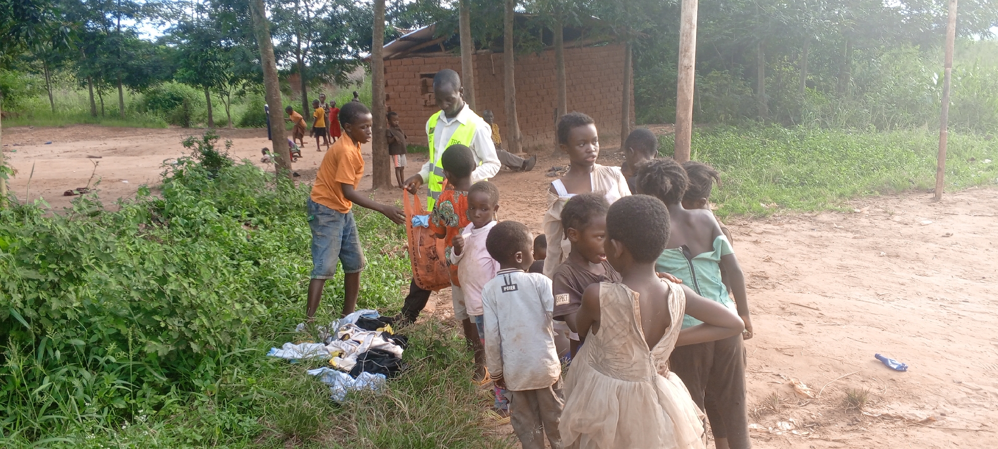 In Ngalula, Democratic Republic of the Congo, a group of children, some in worn clothing, are gathered near a man in a fluorescent vest. A young boy hands clothing from a pile in the grass to the man's open bag. Some children look at the clothes. A brick building is partially visible in the background, along with trees. The ground is dirt.