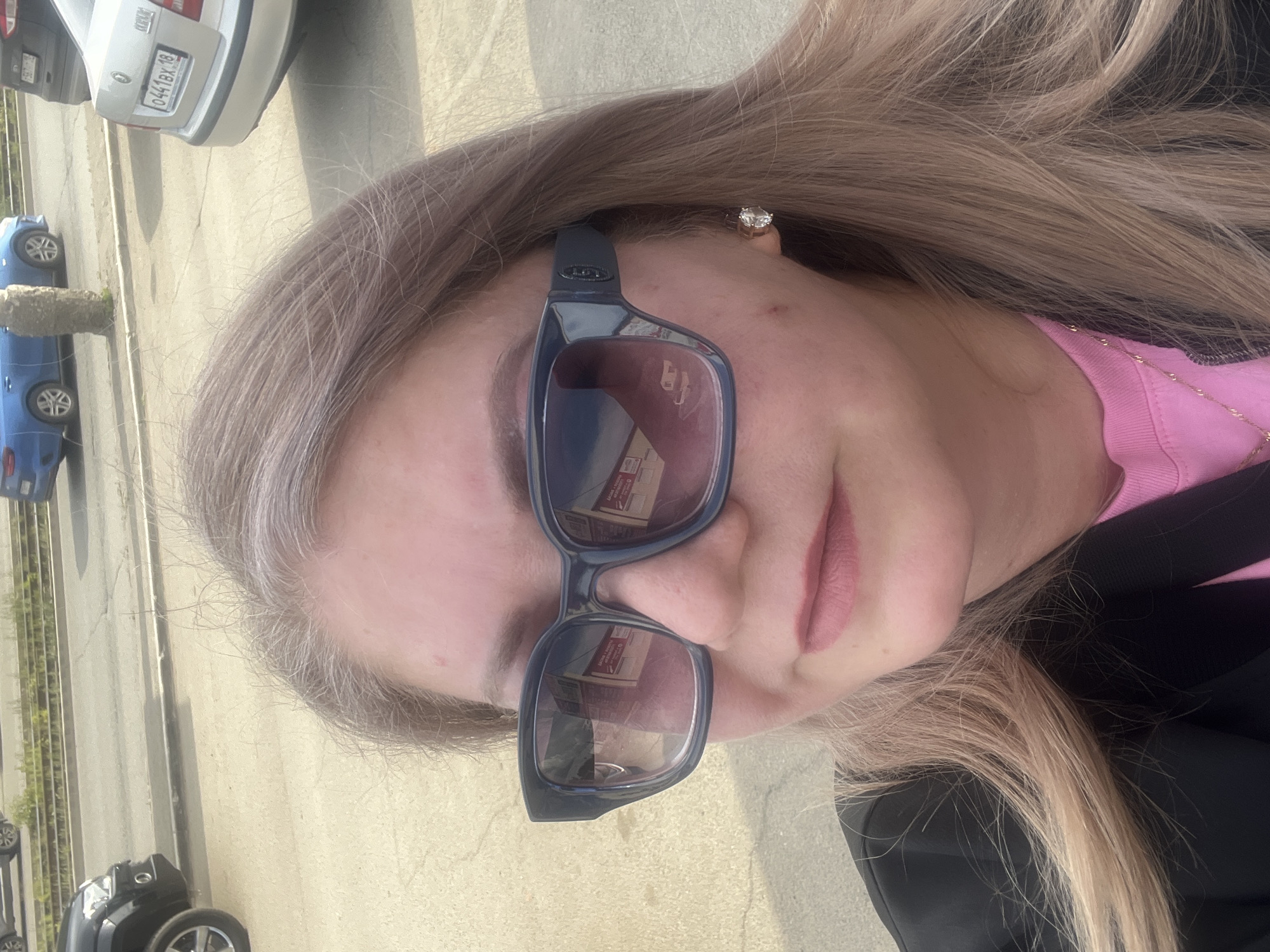 A close-up, rotated portrait captures a woman outdoors on a bright, sunny day, likely in Izhevsk, Russia. She is shown from the chest up, featuring long, wavy, light brown or blonde hair that frames her face. She wears dark-framed sunglasses that cover her eyes, a pink ribbed top, and a black outer garment. A delicate gold-colored necklace is visible around her neck, and a sparkling stud earring adorns her right ear. Her facial expression appears neutral, with her lips slightly parted.
The background reveals an urban or suburban street scene. A paved road or parking lot is visible, with a curb or divider and some sparse greenery. Several cars are parked or moving in the distance, including a silver sedan with a visible Russian license plate `0441BX 18`. The '18' code on the plate corresponds to the Udmurt Republic, confirming the provided location context of Izhevsk. Other vehicles include two blue cars and a black SUV or sedan. Indistinct text, possibly Cyrillic, is reflected in the lenses of her sunglasses, suggesting nearby signage or buildings. The overall scene indicates clear weather during daylight hours.